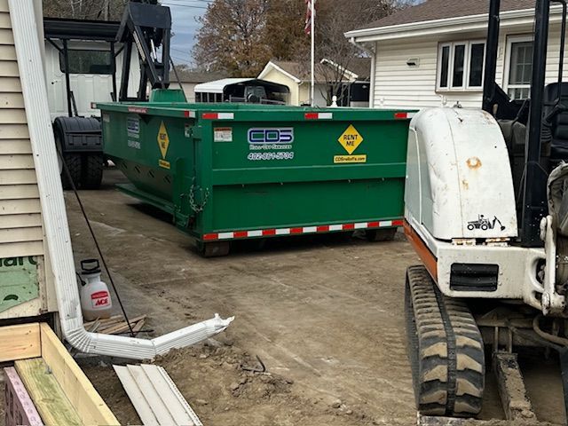 A bright green roll-off dumpster sits in a dirt driveway next to an excavator and a residential house.