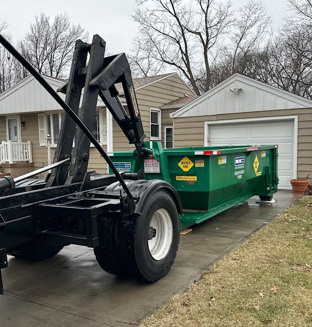 A roll-off truck is positioned on a residential driveway, preparing to drop off a large, bright green waste dumpster.