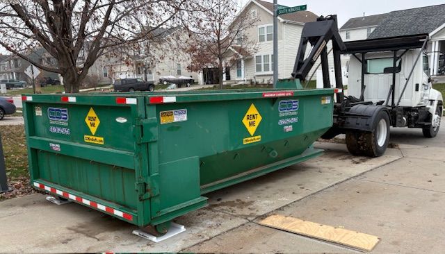 A green dumpster sits on a residential driveway, connected to a white hook-lift roll-off truck in a suburban neighborhood.