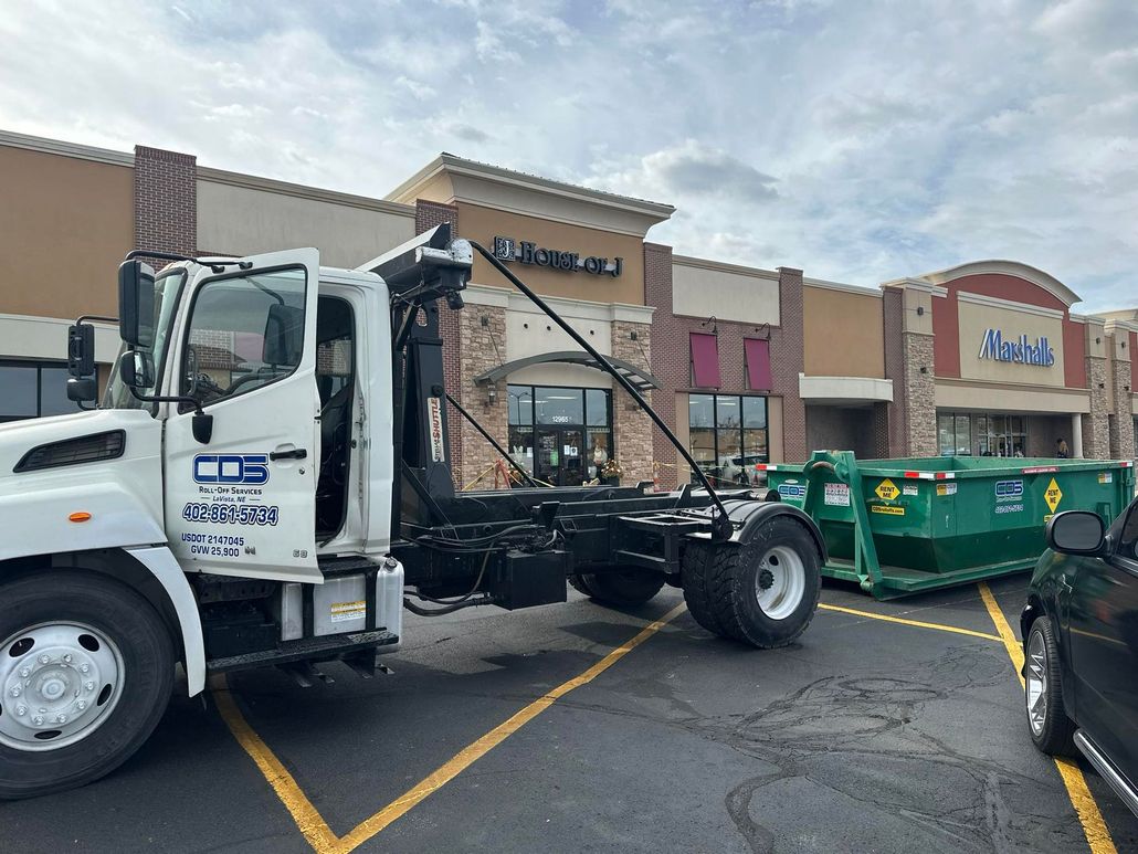 A green dumpster sits on a residential driveway, connected to a white hook-lift roll-off truck in a suburban neighborhood.
