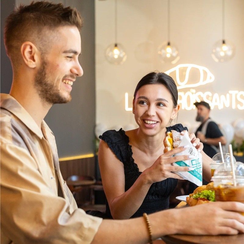 A man and a woman are sitting at a table in front of a sign that says croissants.