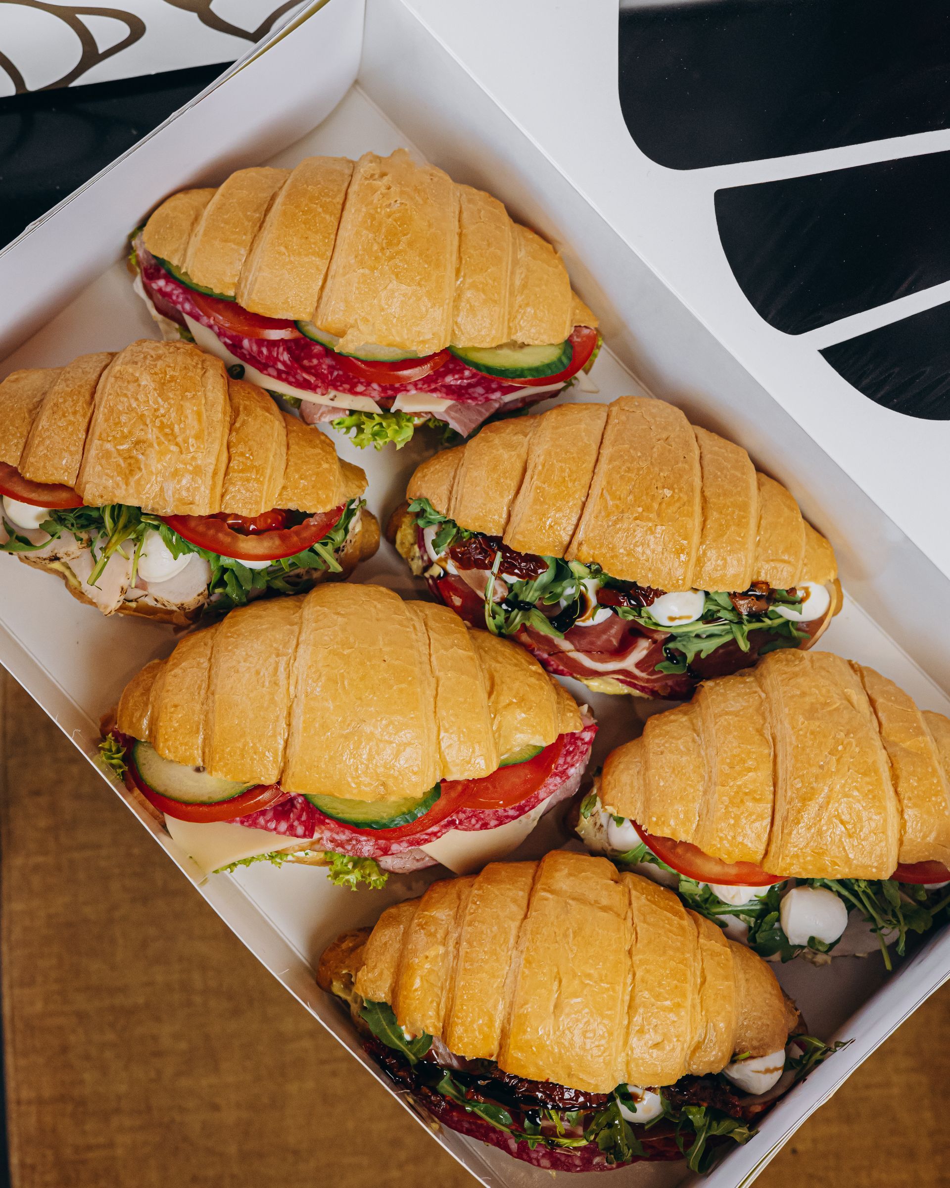 Six croissant sandwiches in a white box with various fillings, including meats, tomatoes, and greens.