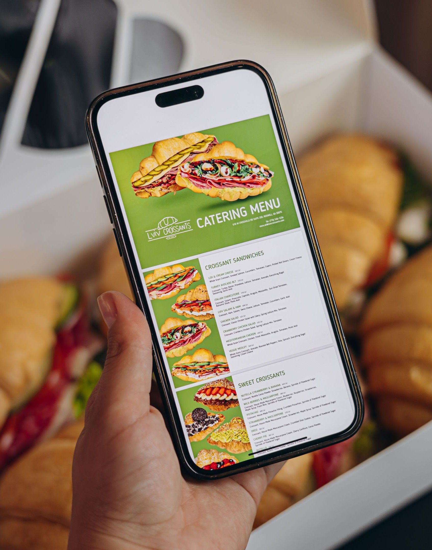 A hand holding a phone displaying a cafe menu, with sandwiches in the background.