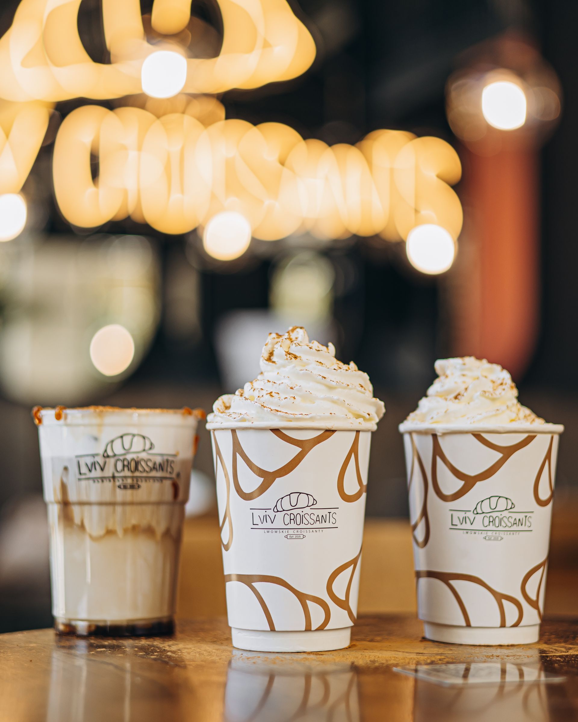 Three beverages with whipped cream and a decorative sign, lit by warm lights.
