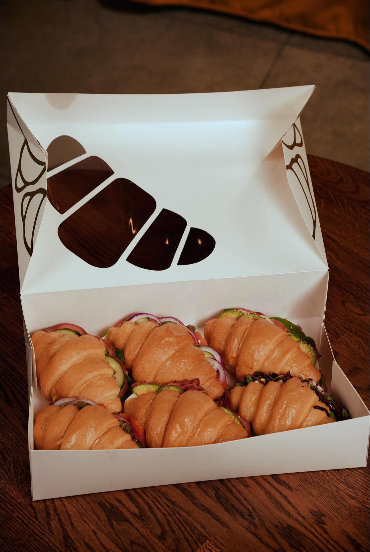 A box filled with croissants and sandwiches on a wooden table.