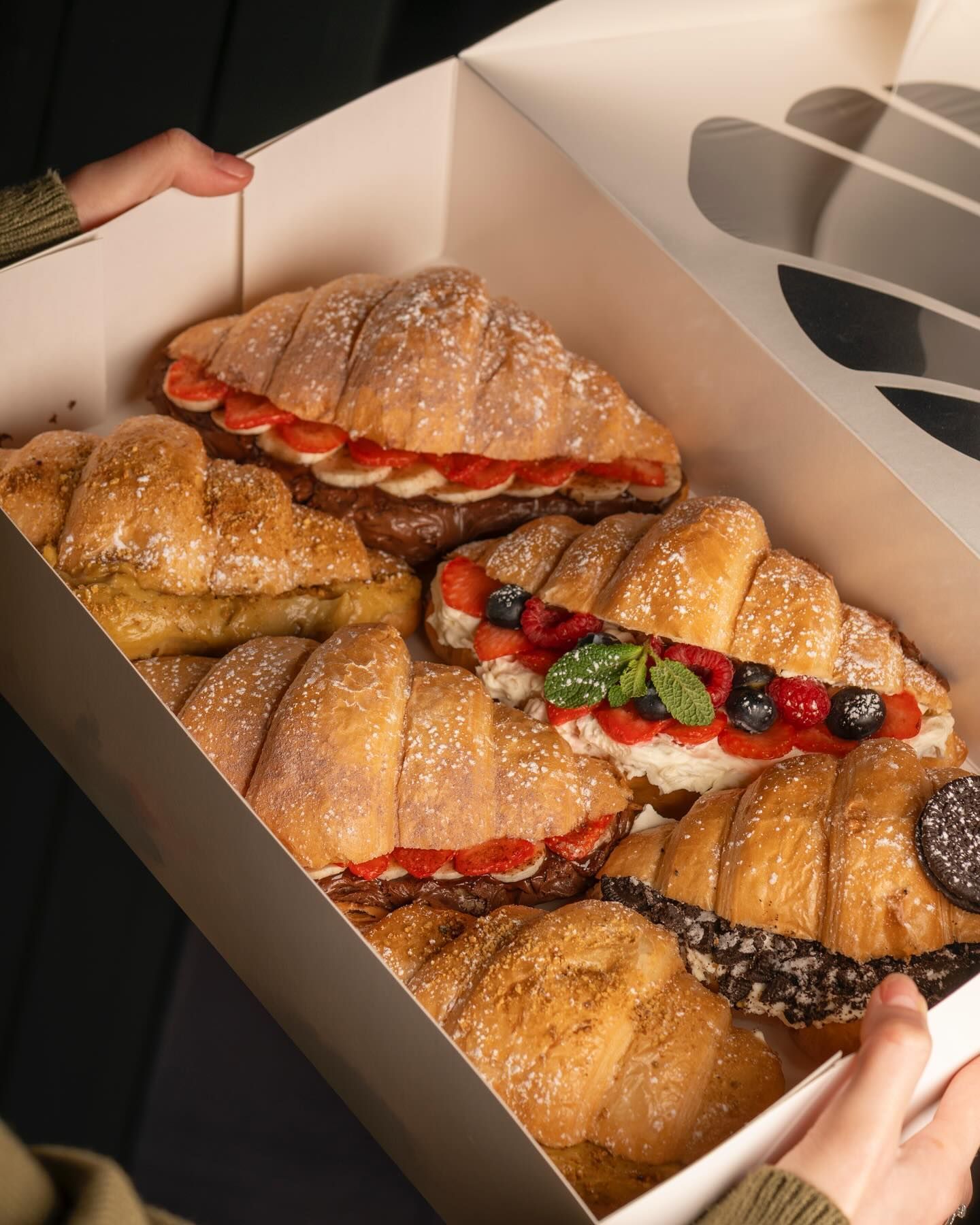 A person is holding a box of croissants with different toppings.