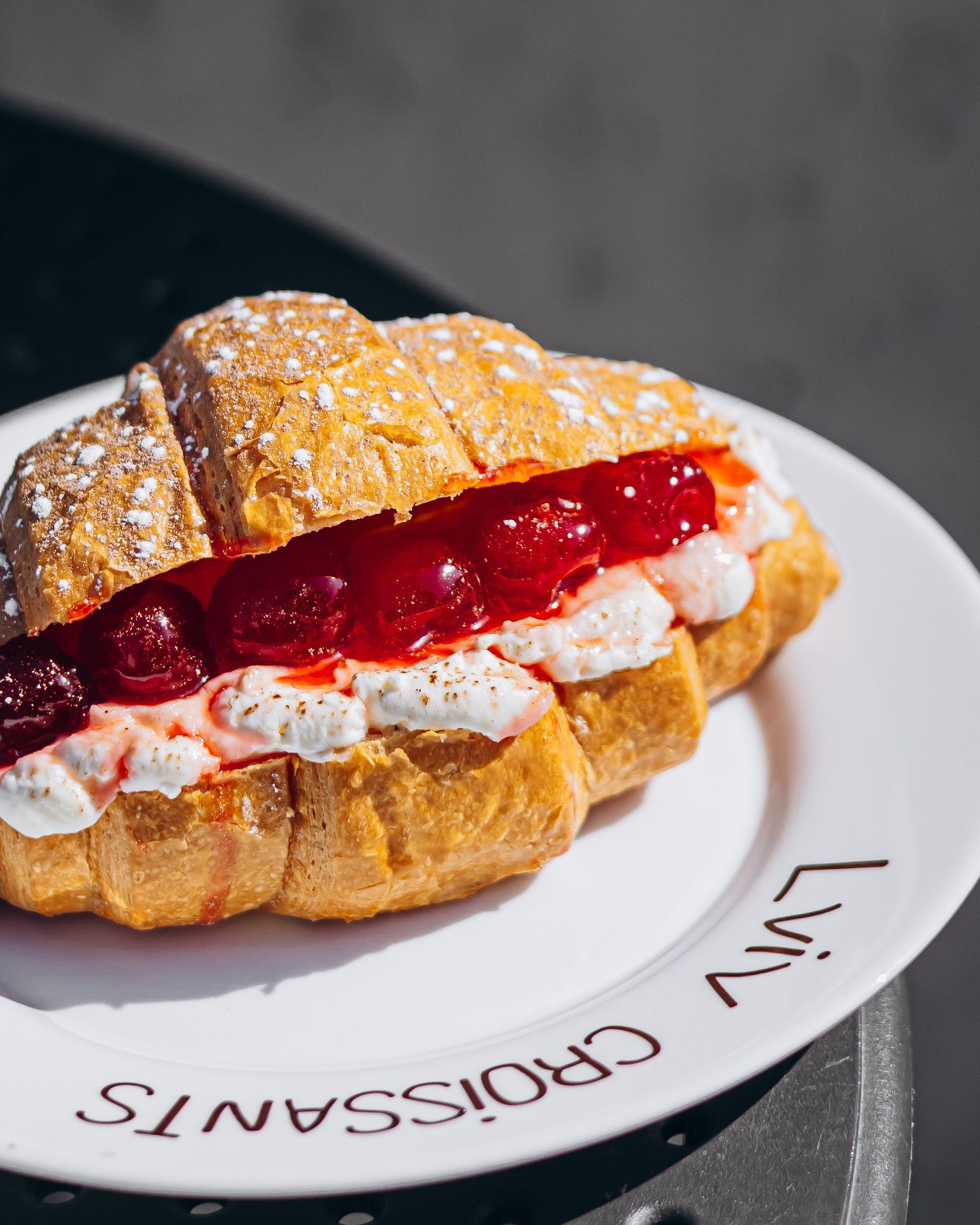 Croissant sandwich with cream and red jam on a white plate, outdoors.