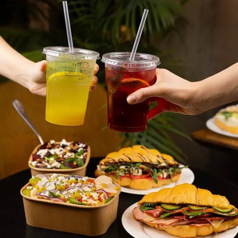 Two people are toasting with drinks and sandwiches on a table.