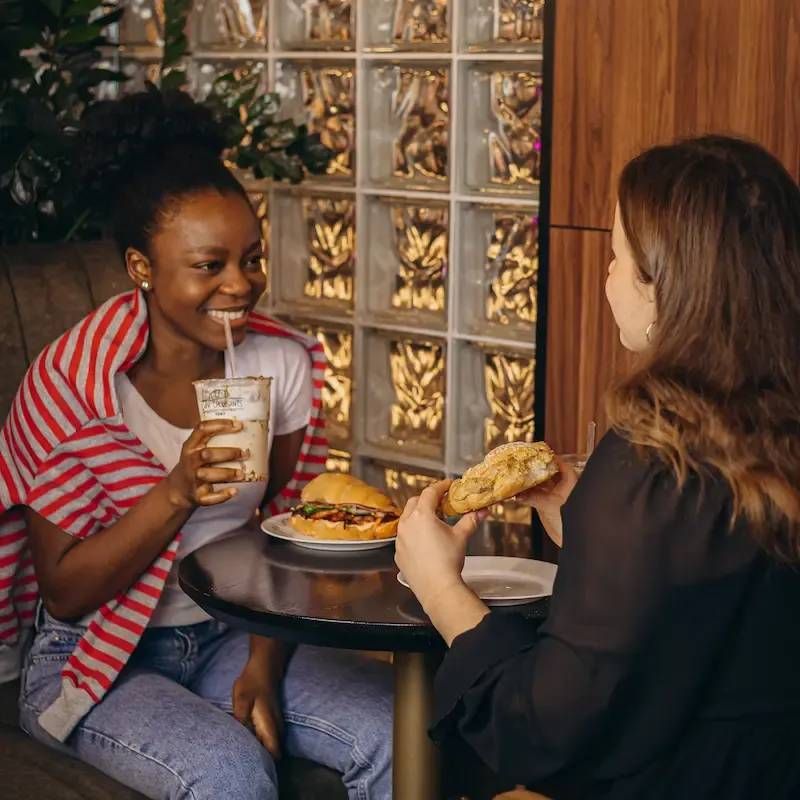 Two women are sitting at a table eating sandwiches and drinking milkshakes.