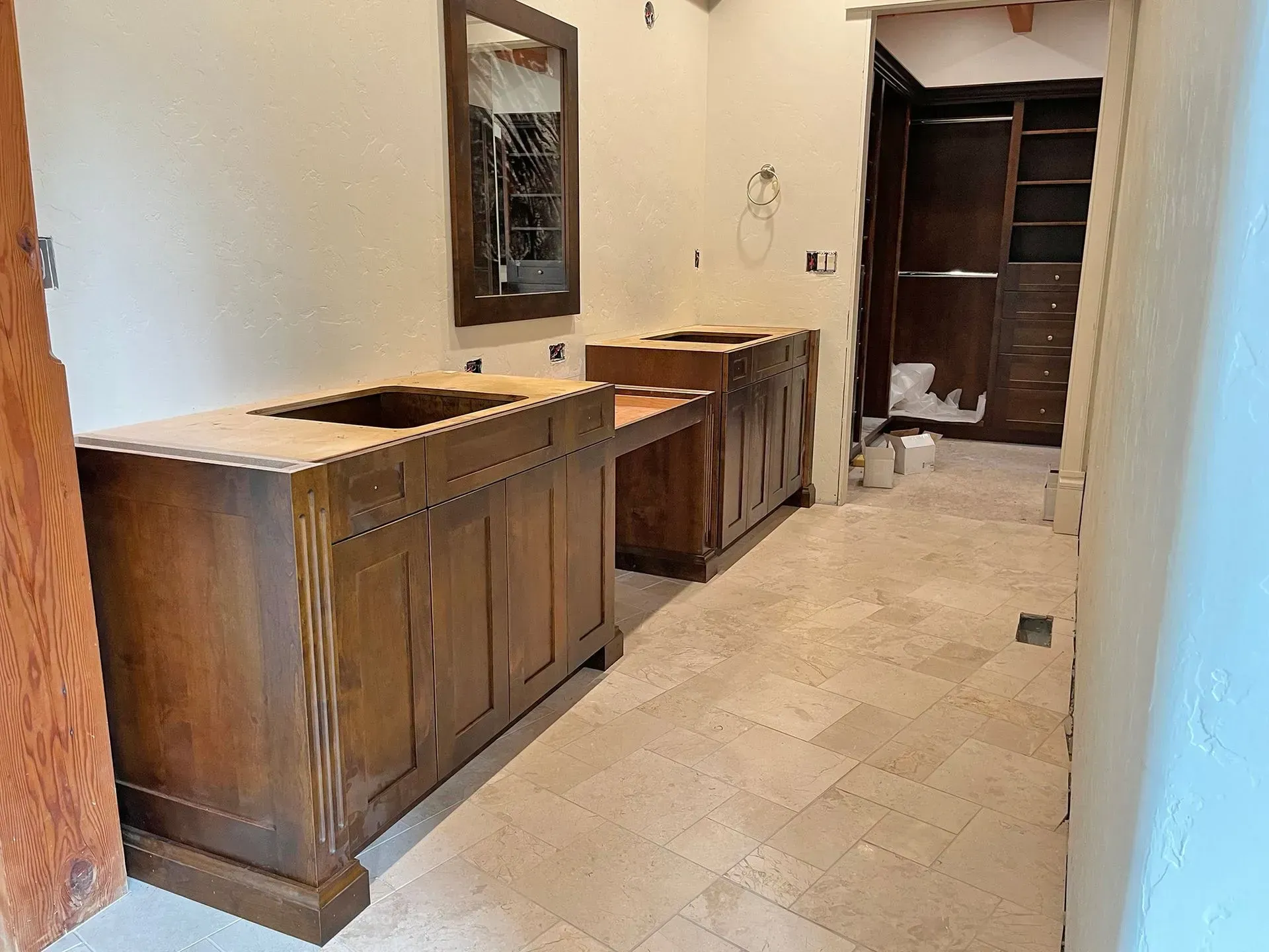 Empty bathroom with wooden vanity cabinets, beige tile floor, and open door to a darker room.