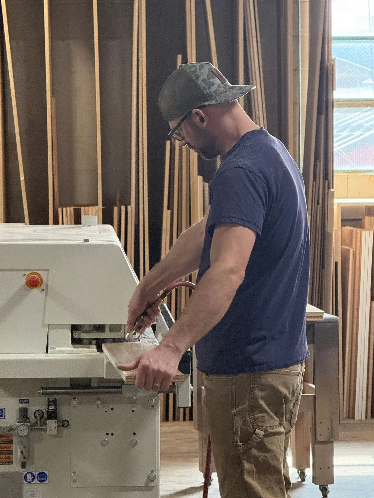 Worker operating a table saw in a woodworking shop, wearing a cap and navy shirt.