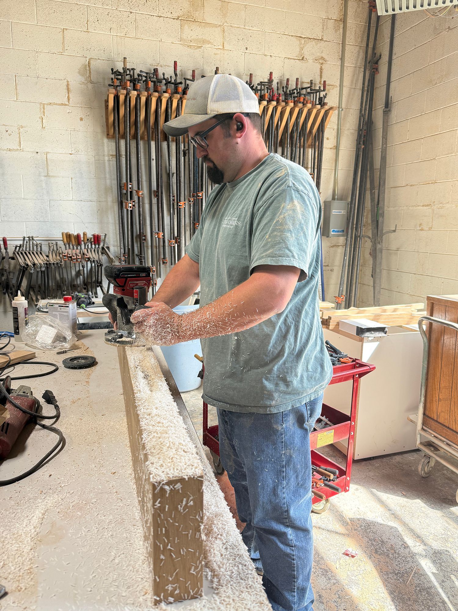 Worker sanding a wooden board in a workshop, surrounded by tools and sawdust.