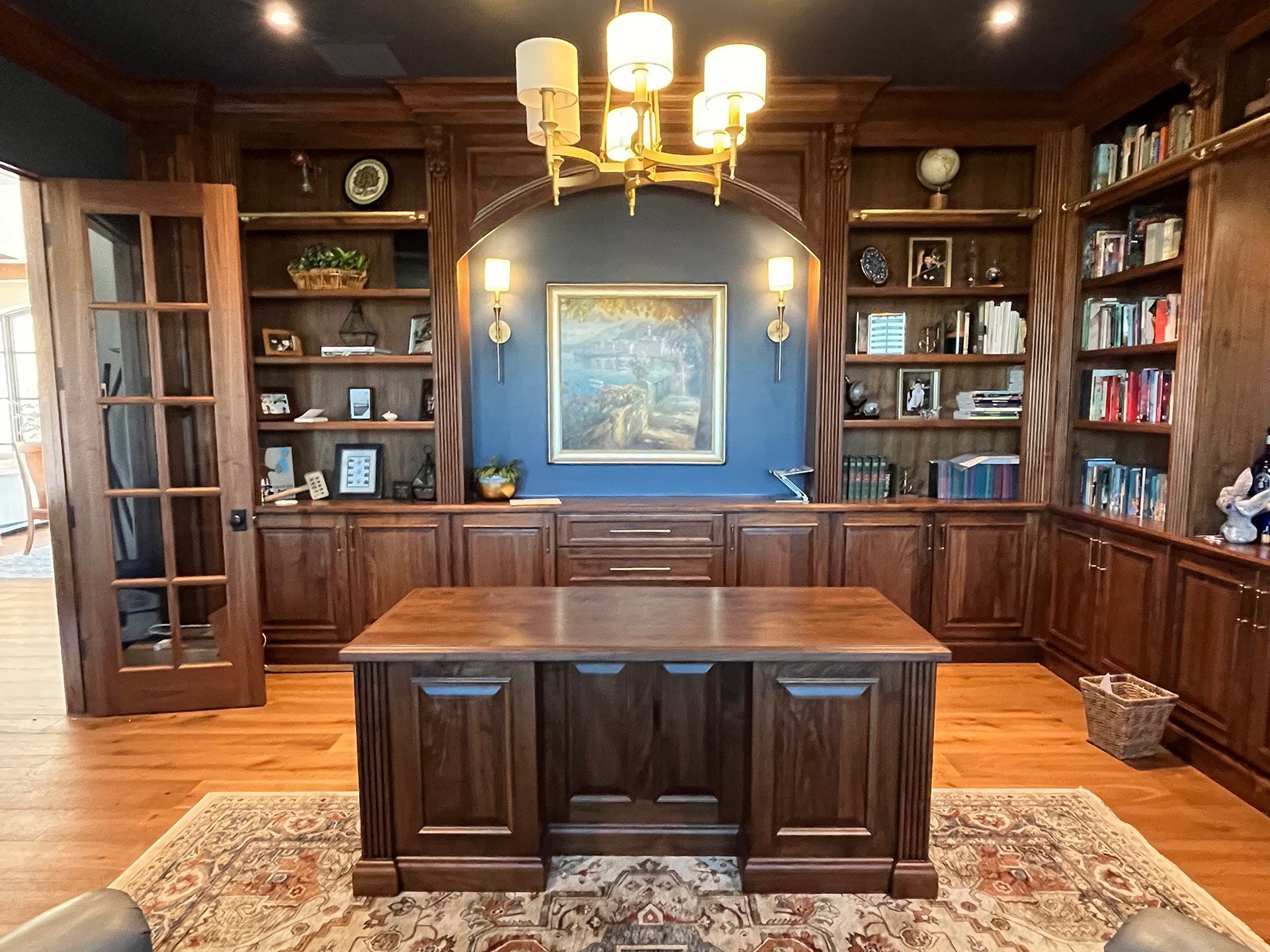 Elegant wood-paneled library with built-in bookshelves, central desk, chandelier, and framed art on a blue wall