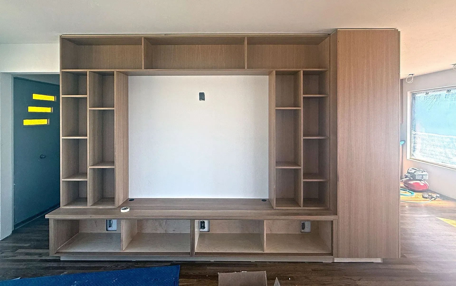 Built-in wooden wall unit with shelves, cabinets, and a large central TV opening in a room under renovation.