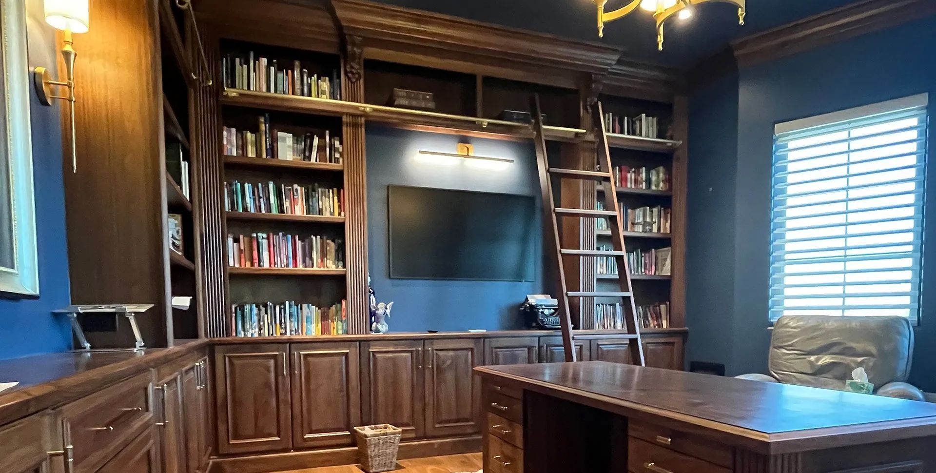 Wood-paneled home office with bookshelves, large TV, ladder, and a desk by a window with blinds.