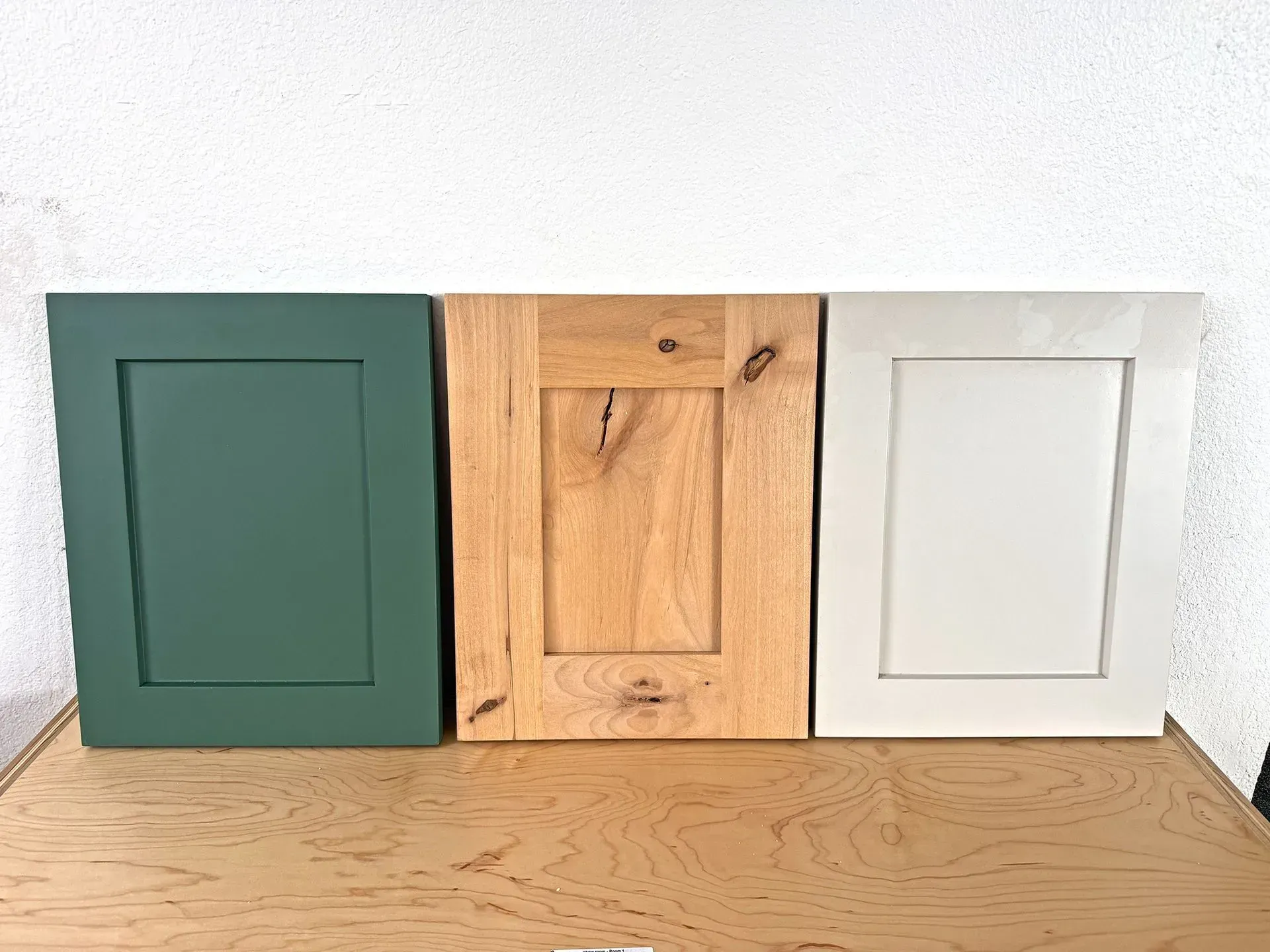 Three decorative square wall panels in green, wood, and white leaning against a wall on a wooden surface.