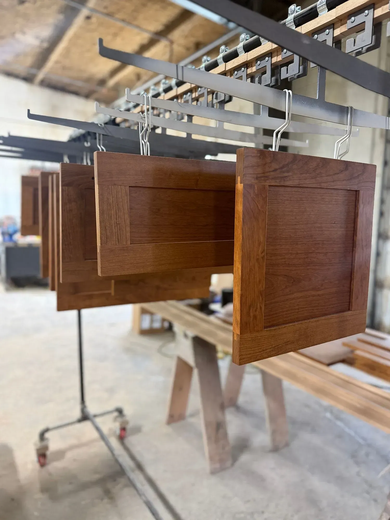 Wooden cabinet doors hanging on a workshop drying rack, with unfinished furniture in the background.