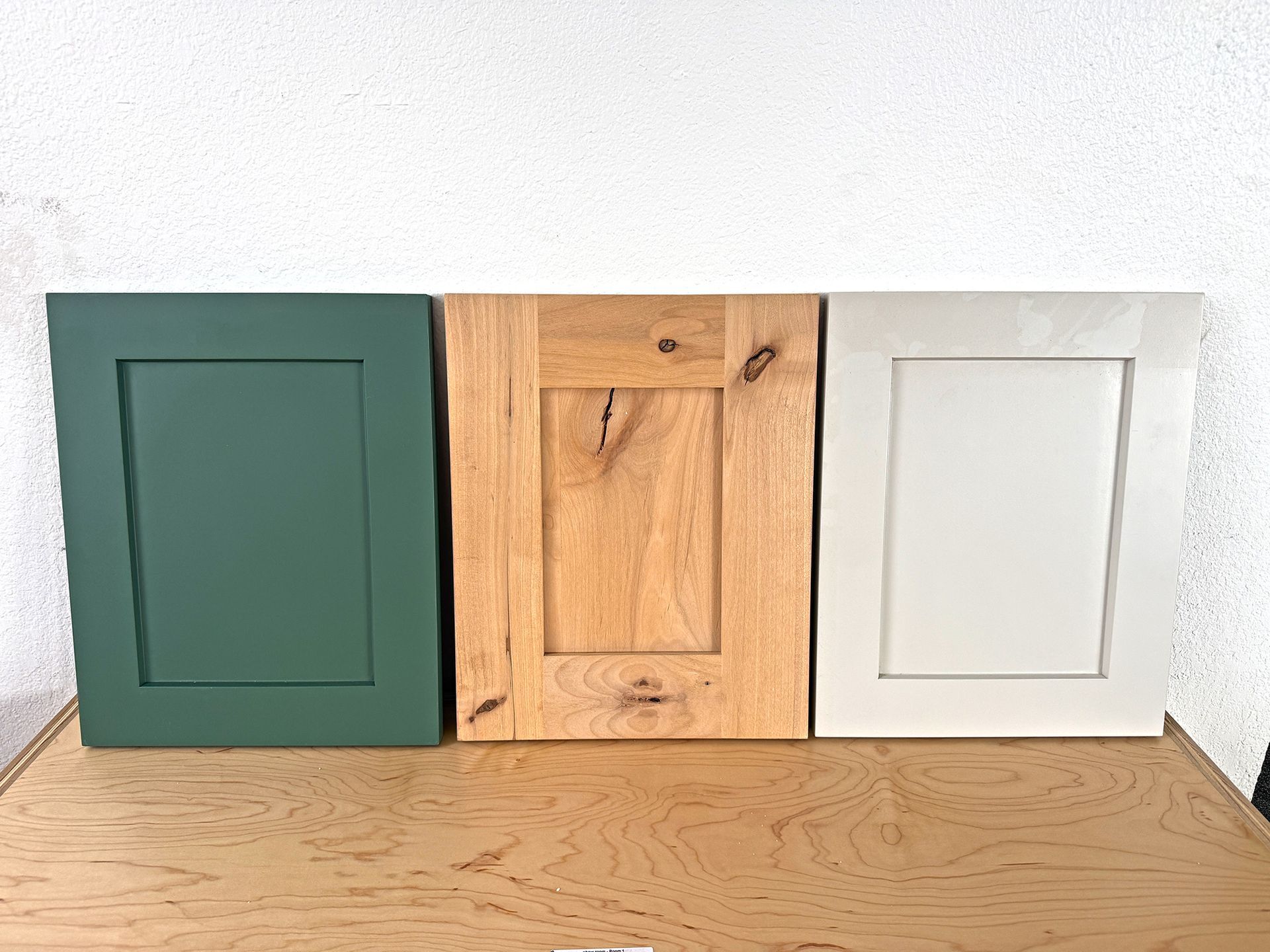 Three square wooden cabinet doors in green, natural wood, and white on a shelf against a textured wall.