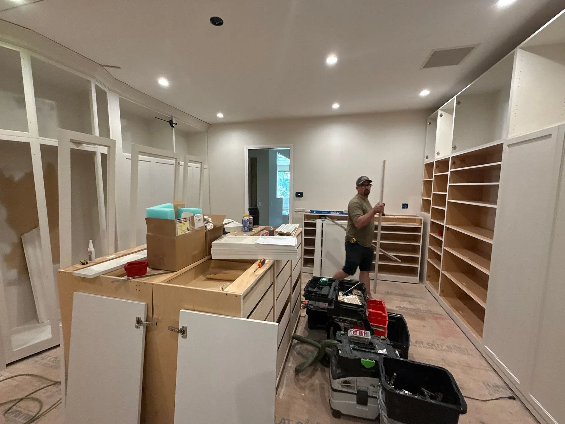 Person standing in a room under renovation with cabinets, tools, and shelves along the walls.
