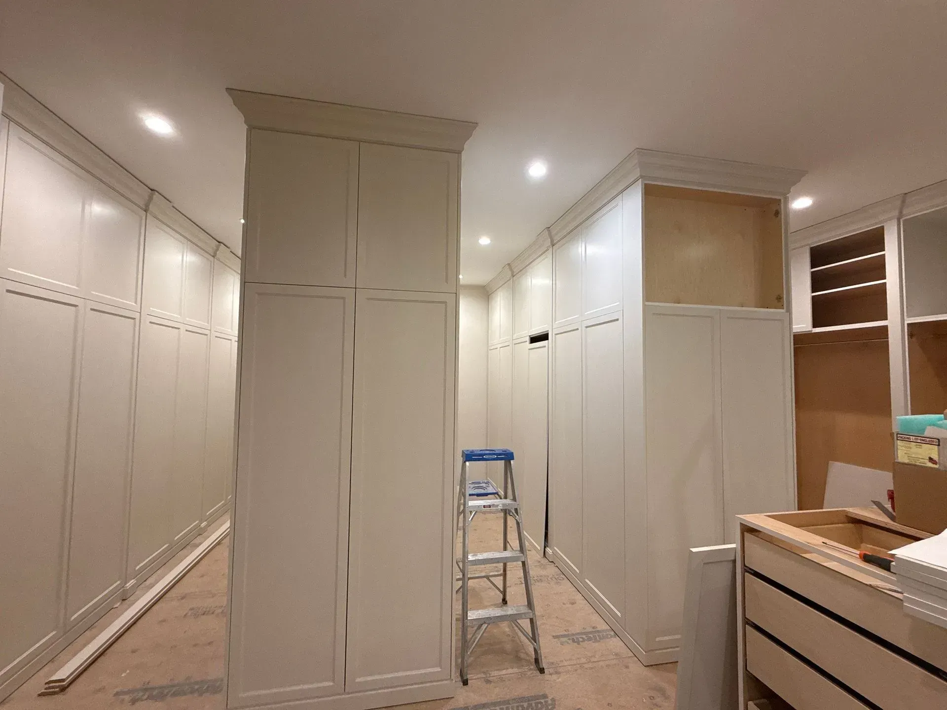 Narrow room under renovation with white built-in cabinets, a ladder, and unfinished wood trim.