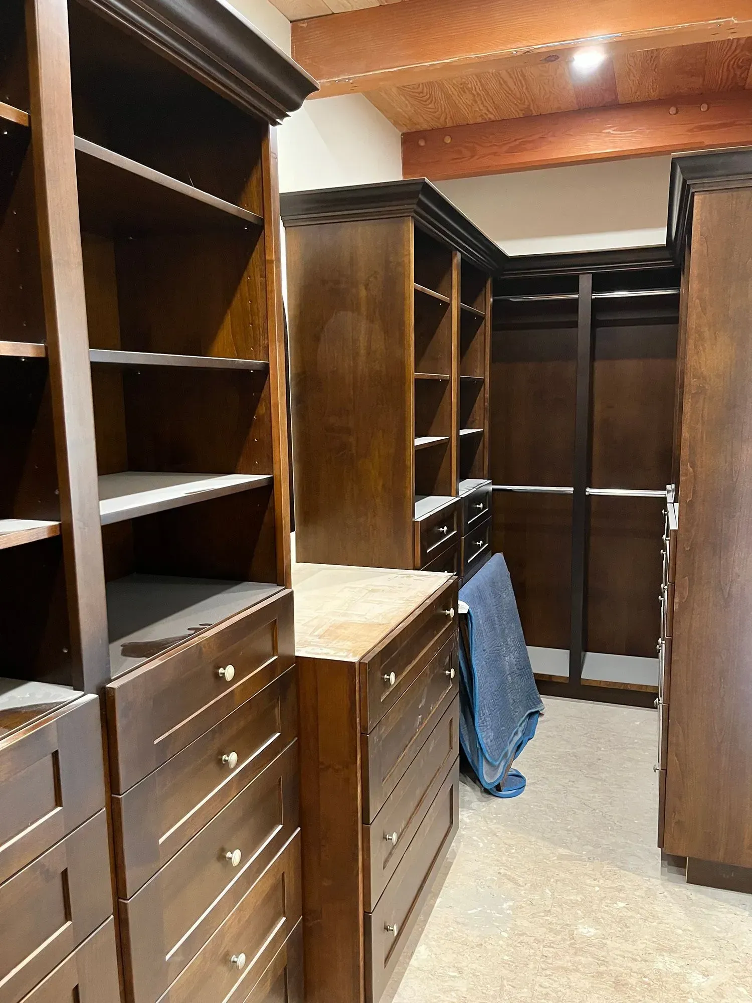 Empty walk-in closet with dark wood shelves, drawers, and hanging rods, lit by warm ceiling lights.