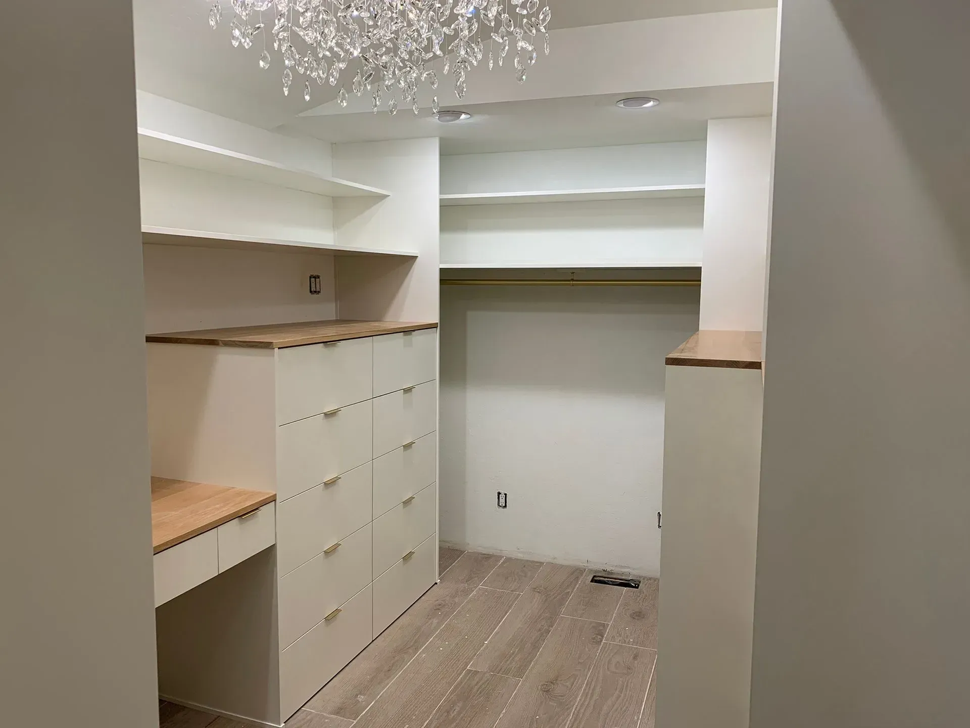 Empty walk-in closet with white shelves, drawers, and wood trim under a crystal light fixture.