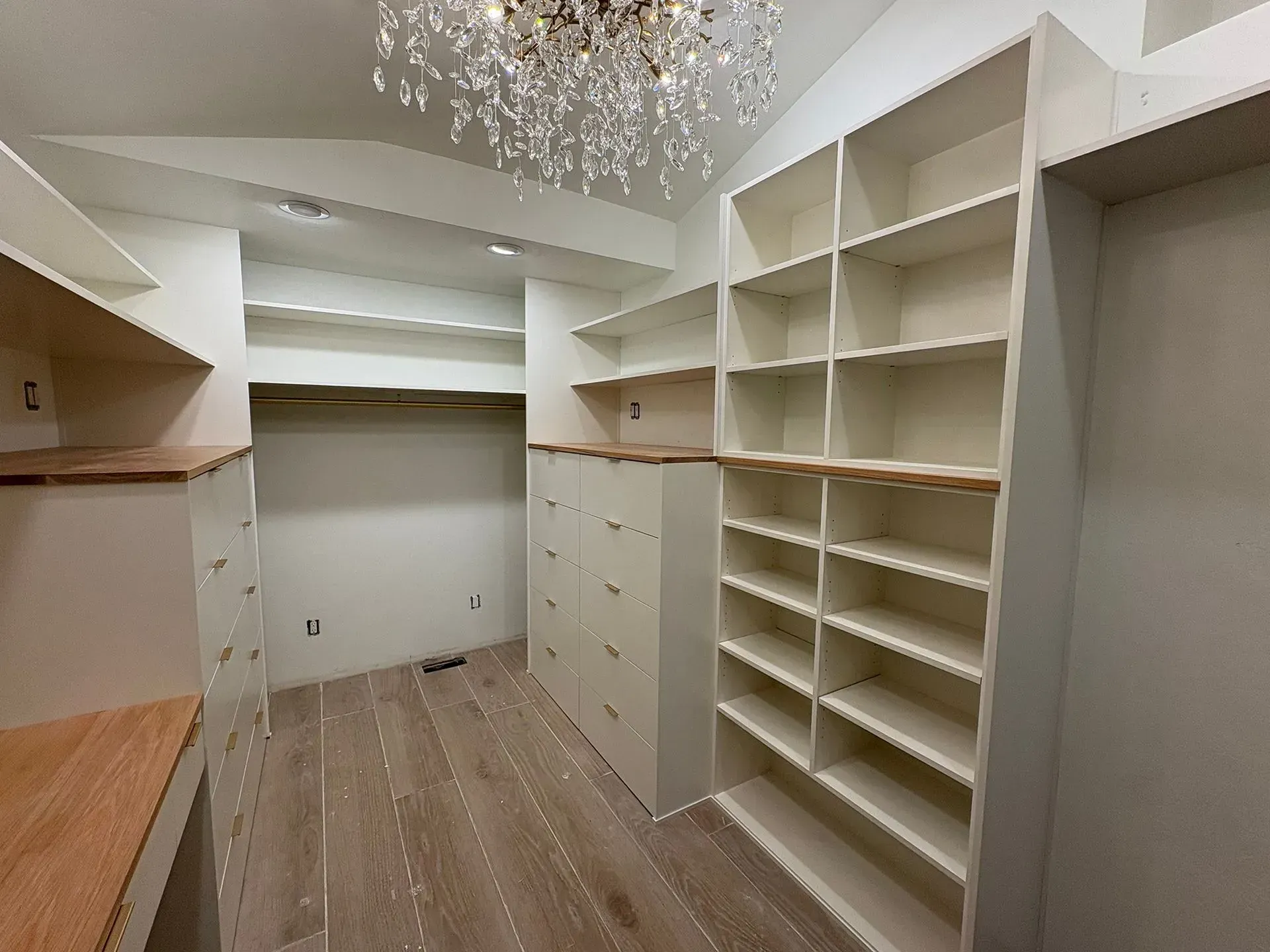 Empty walk-in closet with built-in white shelving and drawers, wood floor, and chandelier overhead.