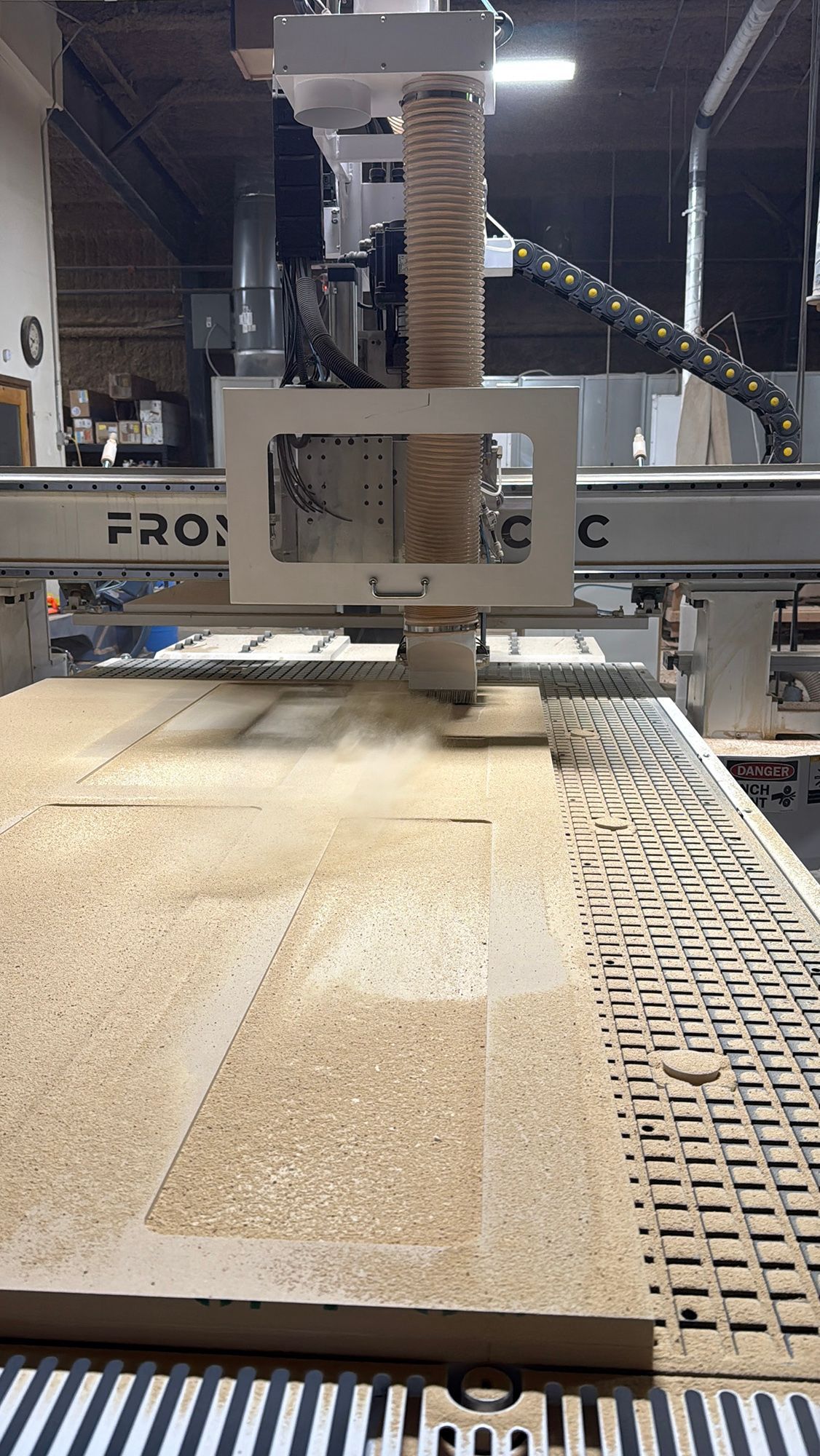 CNC router cutting a light wooden board on a workshop table with dust extraction hose above