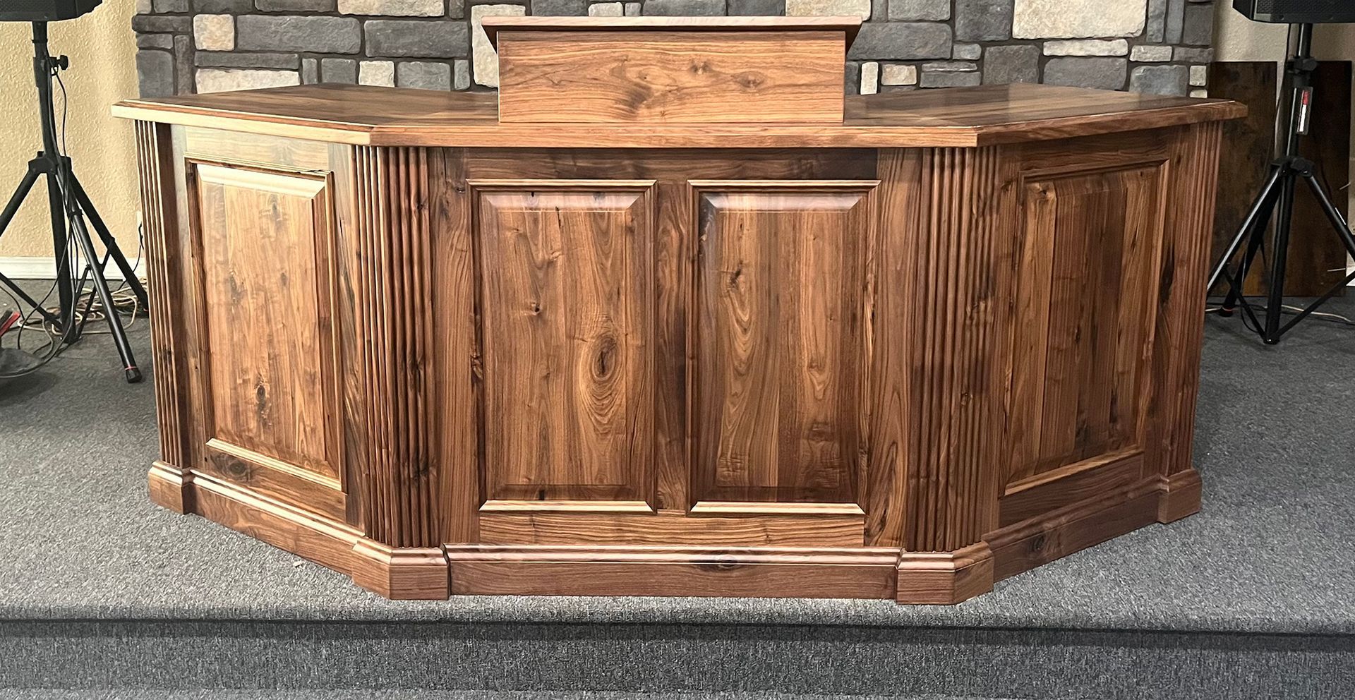 Wooden church pulpit with paneled front, centered on a carpeted stage before a stone wall.