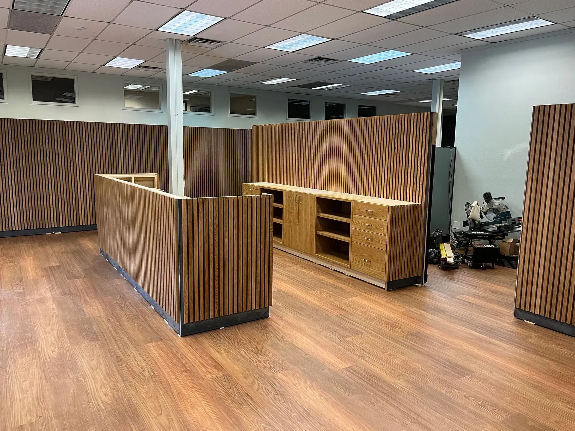Modern office lobby with wood floors, striped wood partitions, and a person seated at a desk in the back right.