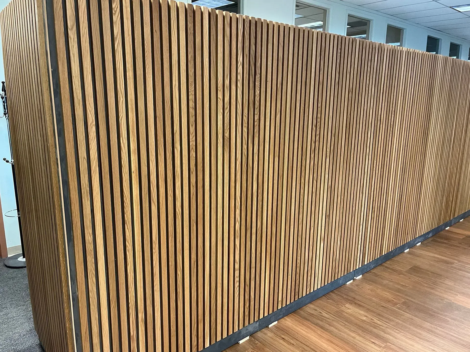 Curved wooden slat wall with black base in a modern indoor space, beside a wood floor.