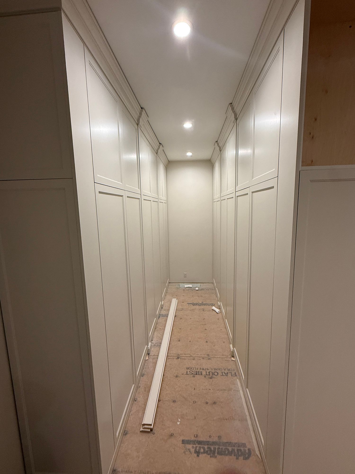 Narrow hallway with white cabinets on both sides and a cardboard-covered floor under recessed lights