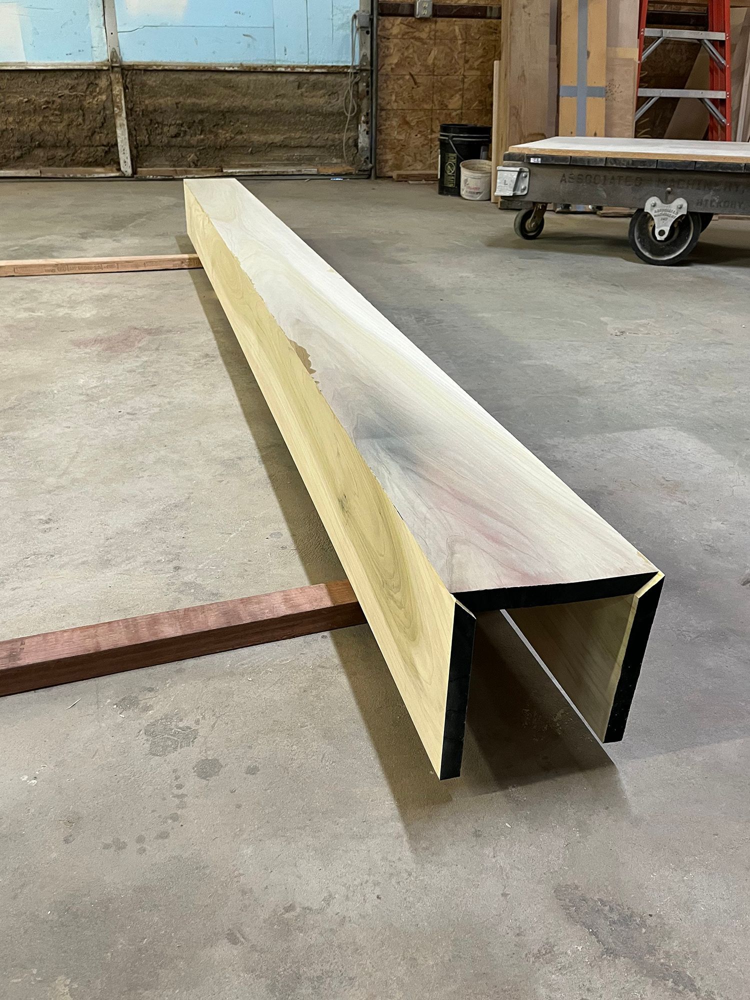 Long steel beam on concrete floor in an industrial workshop, with a cart in the background.