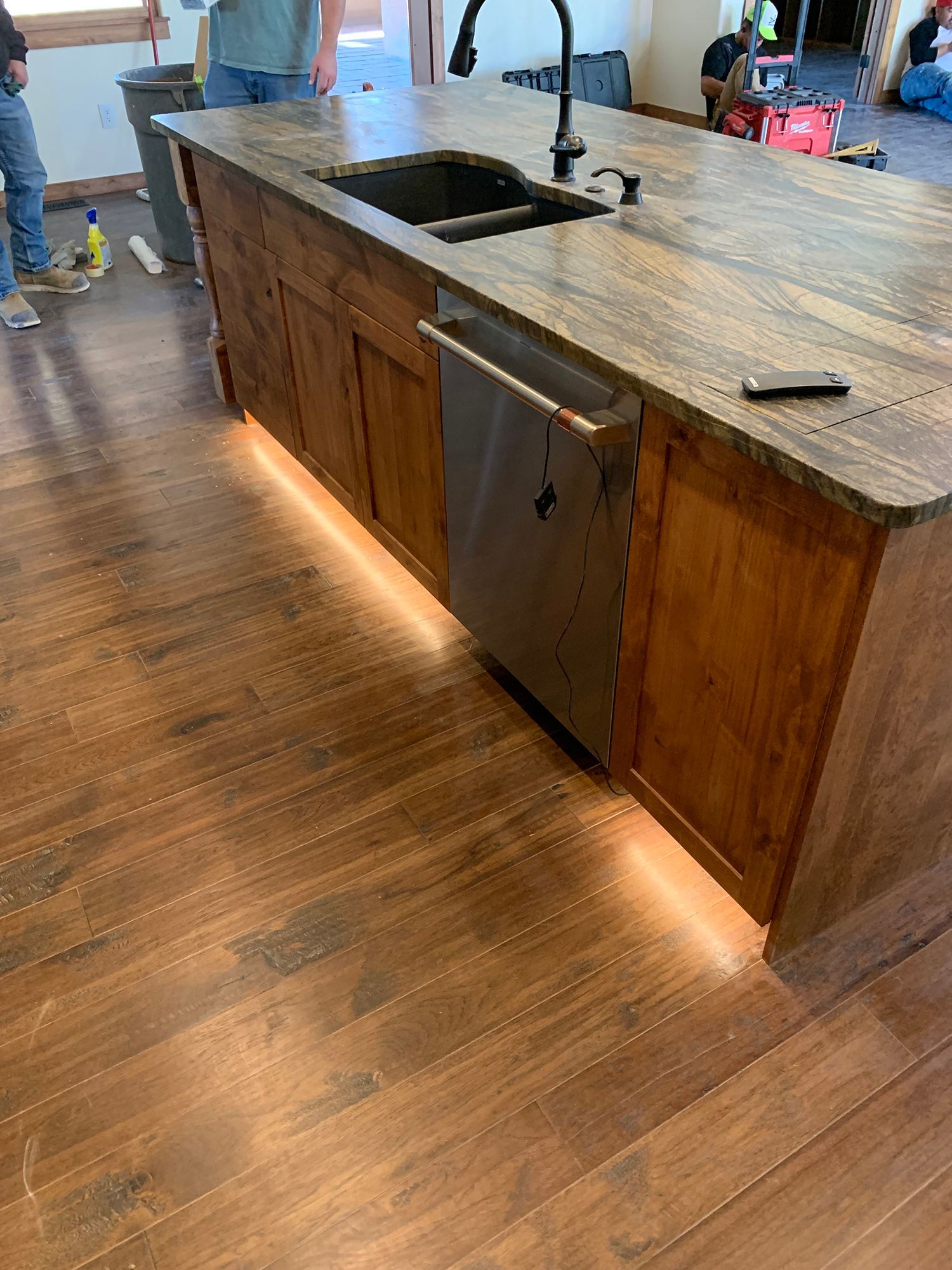 Kitchen island with sink and wood cabinets, lit by under-cabinet lights, in a room with hardwood floor