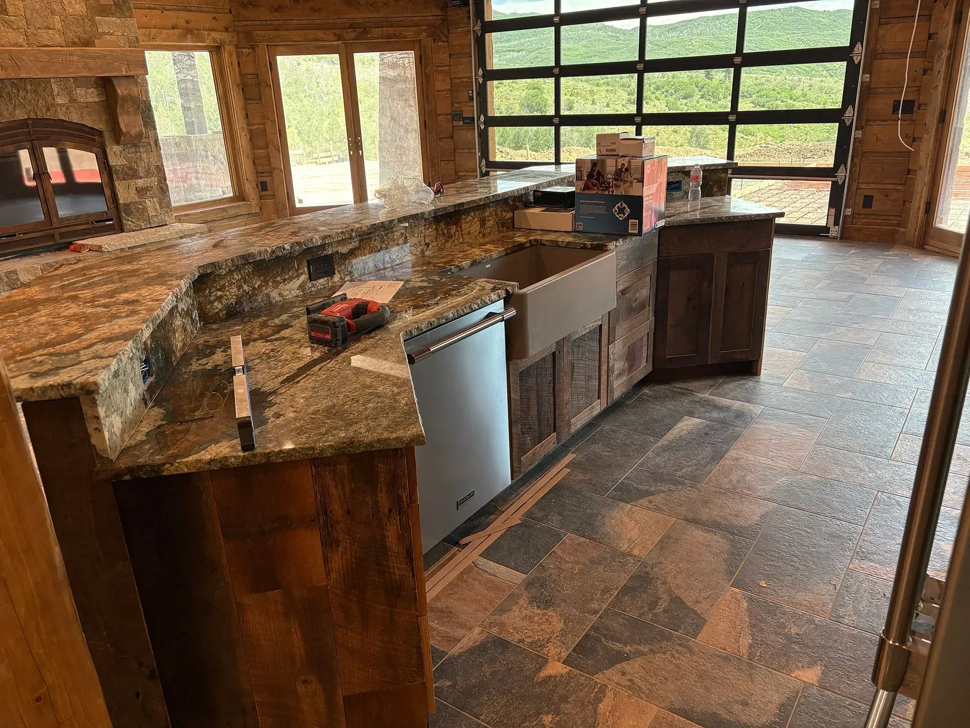 Rustic kitchen with stone island, wood cabinets, and large windows overlooking greenery.