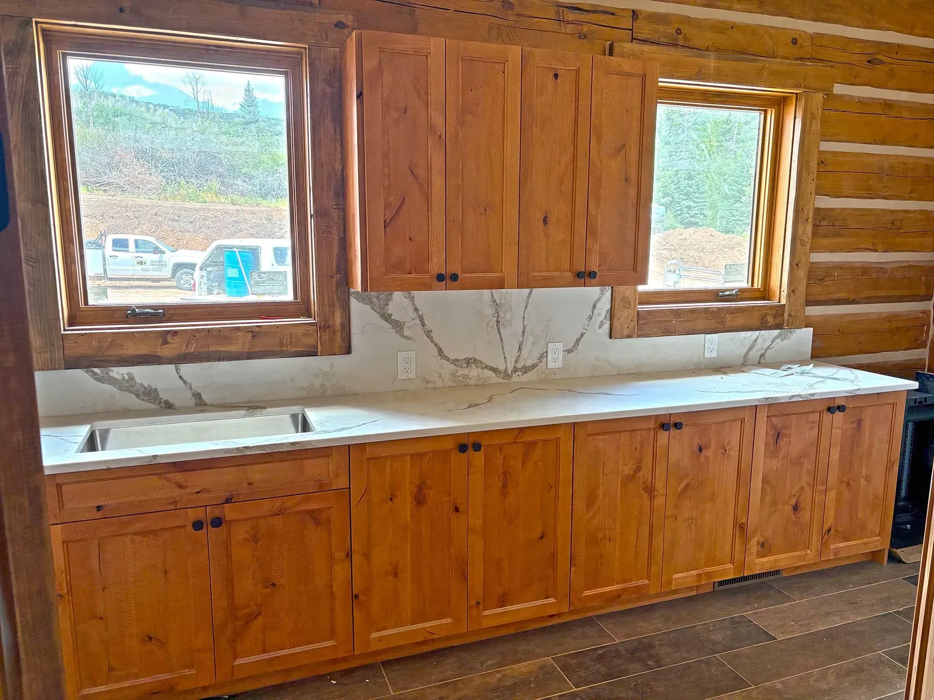 Wooden kitchen cabinets with white countertop beneath two windows in a log cabin interior.