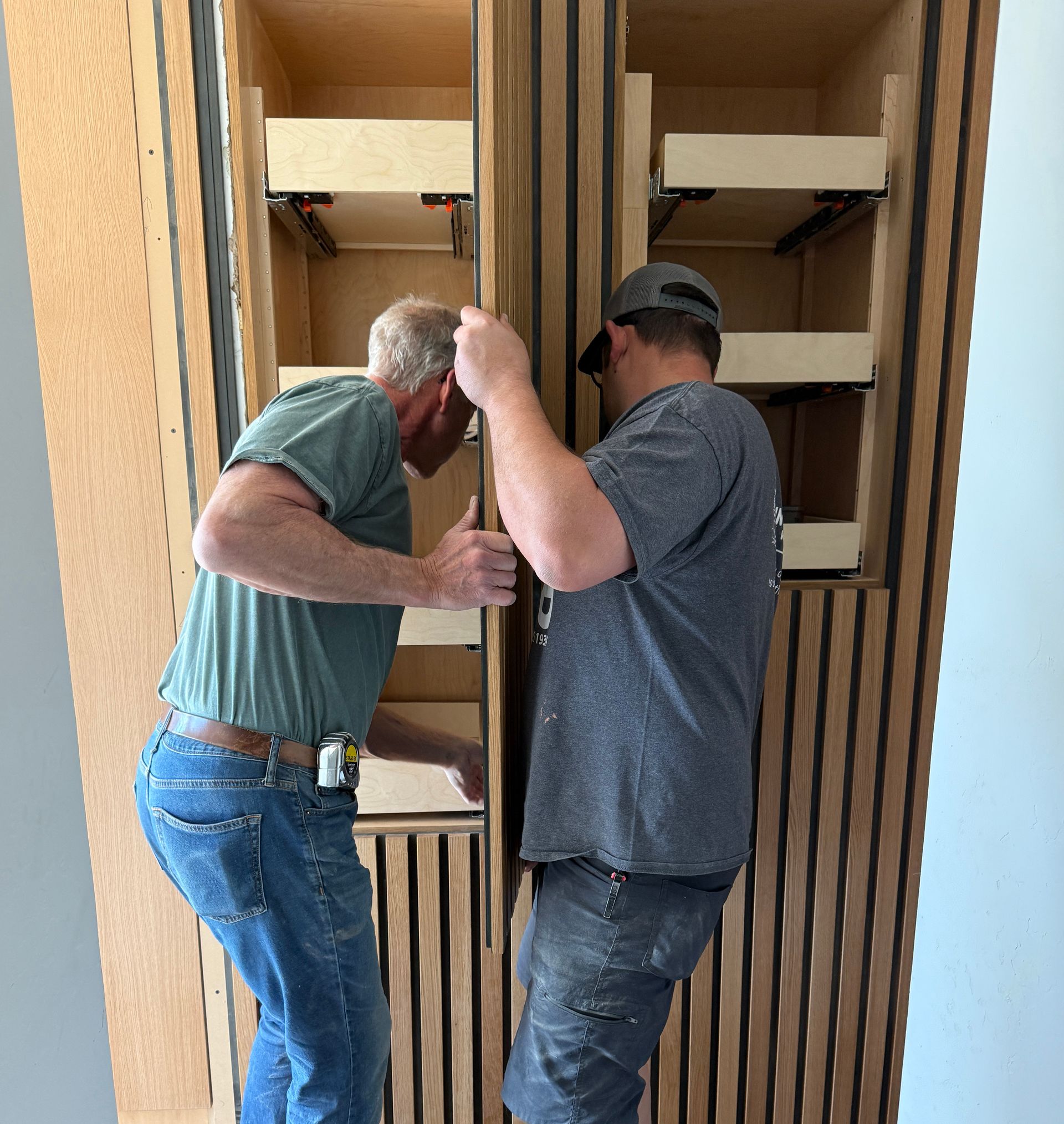Two men assembling a wooden cabinet, one holding a board while the other adjusts a vertical panel.