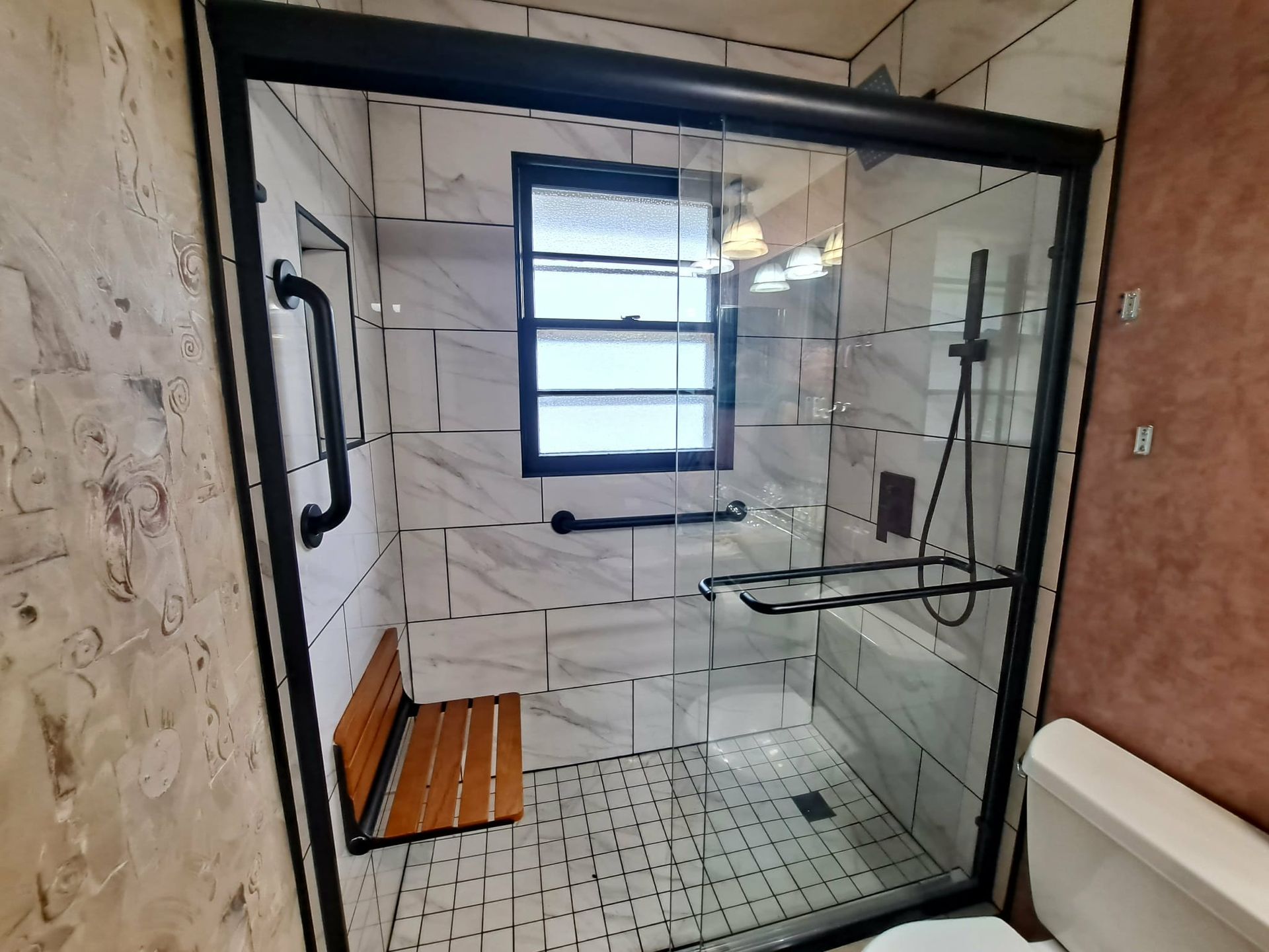 Shower with black frame, glass doors, and grab bars. Features a wooden bench, marble-look tile, and a window.