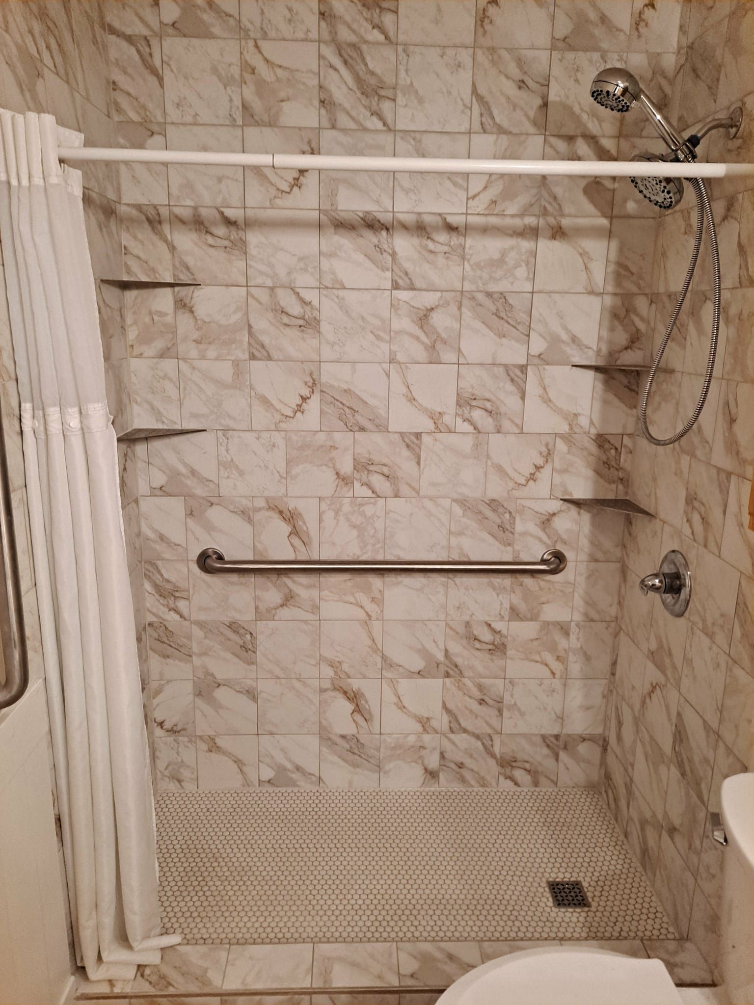 Accessible shower with grab bar, built-in shelves, tiled walls. Shower curtain and shower head visible.