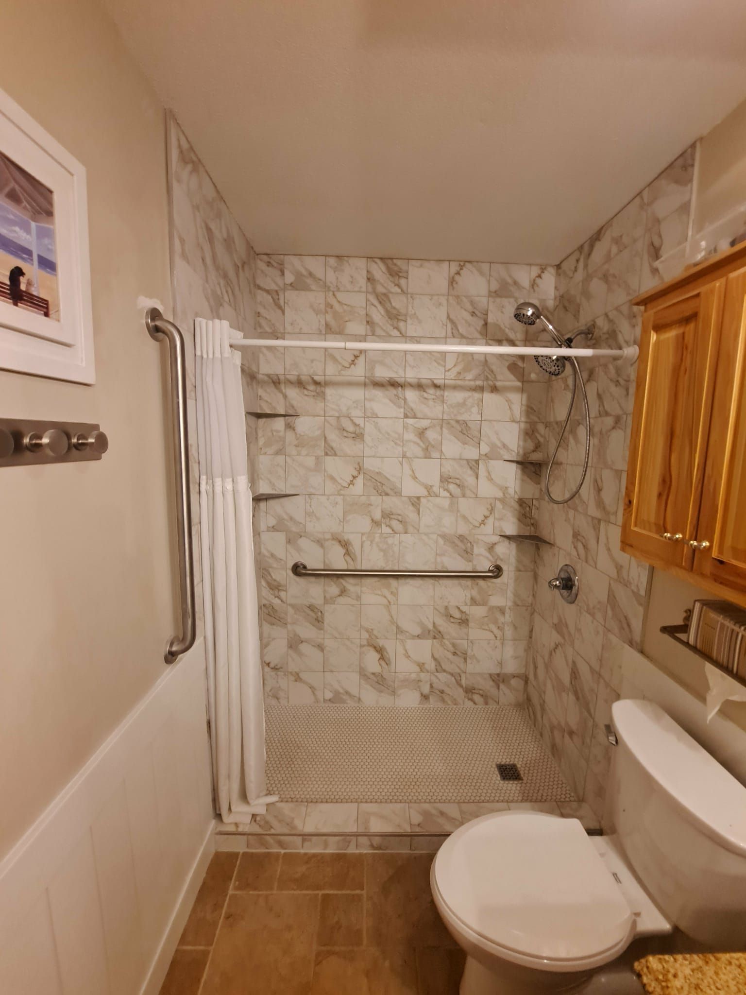 Bathroom with wheelchair-accessible shower. Marble-look tile, grab bars, shower curtain, toilet, and light wood cabinet.