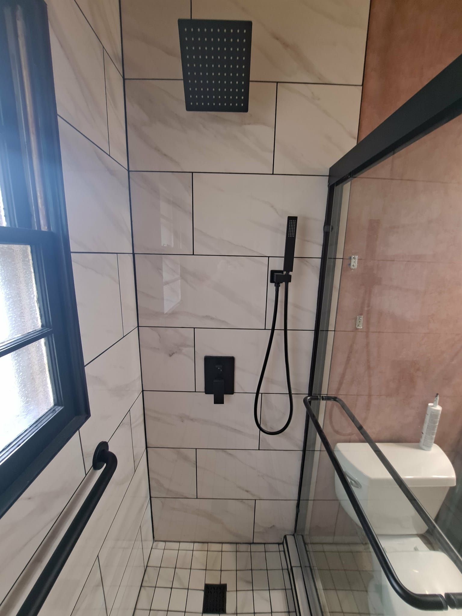Modern shower with black fixtures and white marble-look tiles. Window on the left, glass door on the right.