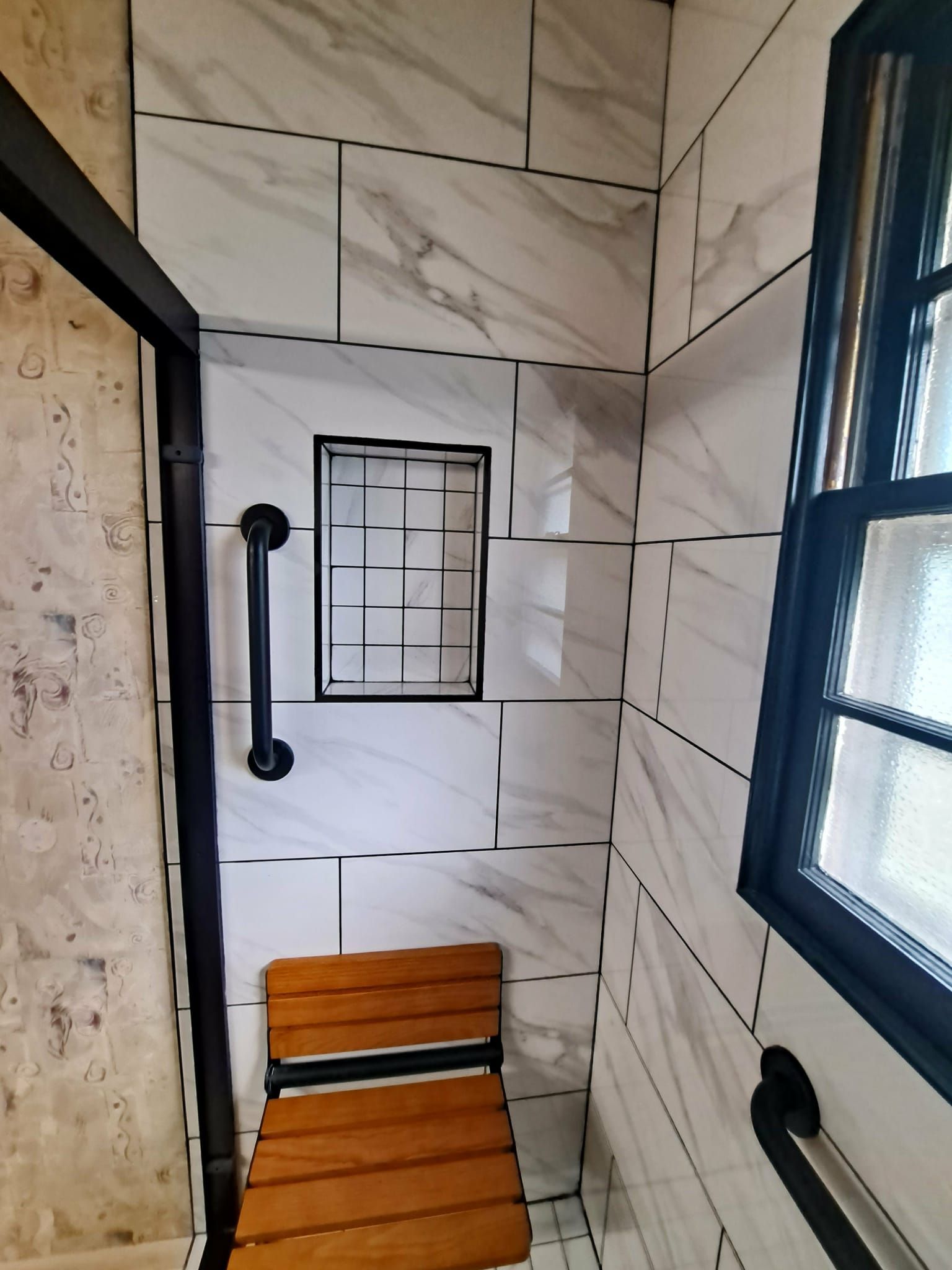 Shower with marble-patterned tiles, a built-in shelf, grab bars, and a wooden fold-down seat.