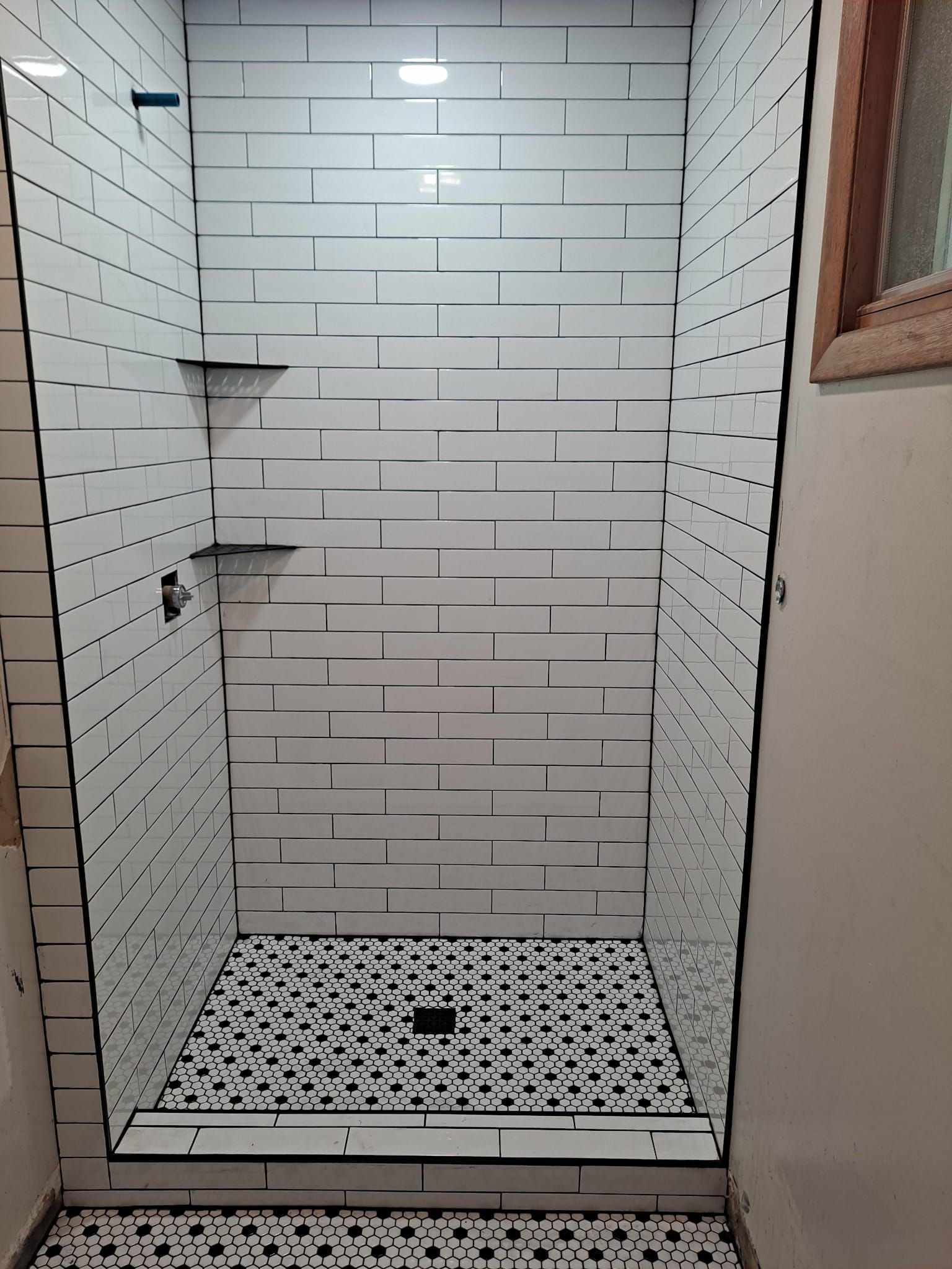 White subway-tiled shower with black grout and black and white patterned floor. Two corner shelves are visible.