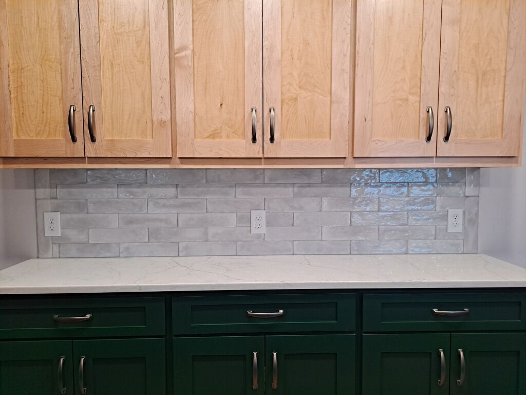 Kitchen with light wood upper cabinets, green lower cabinets, and gray brick tile backsplash.