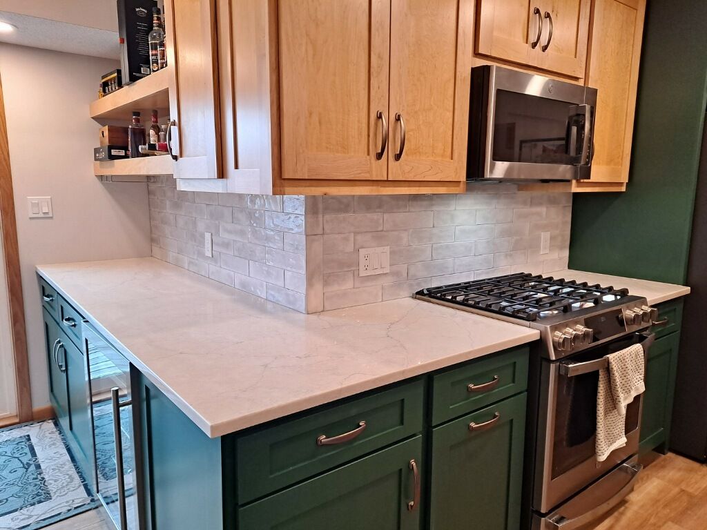 Kitchen with green cabinets, light countertops, and light wood upper cabinets. Backsplash is a light brick pattern.
