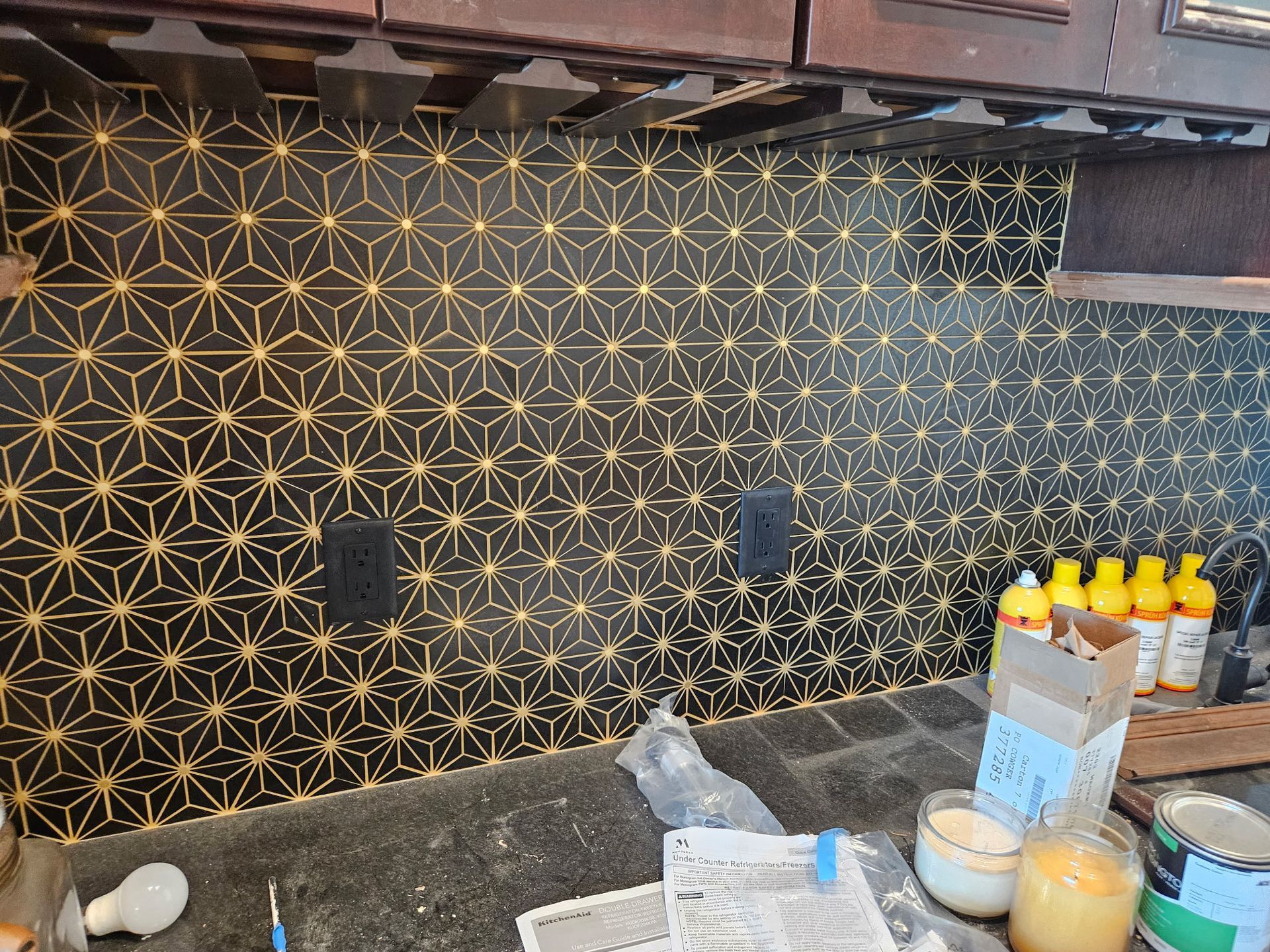 Black and gold geometric tile backsplash installed in a kitchen. Electrical outlets and various tools are visible on the countertop.