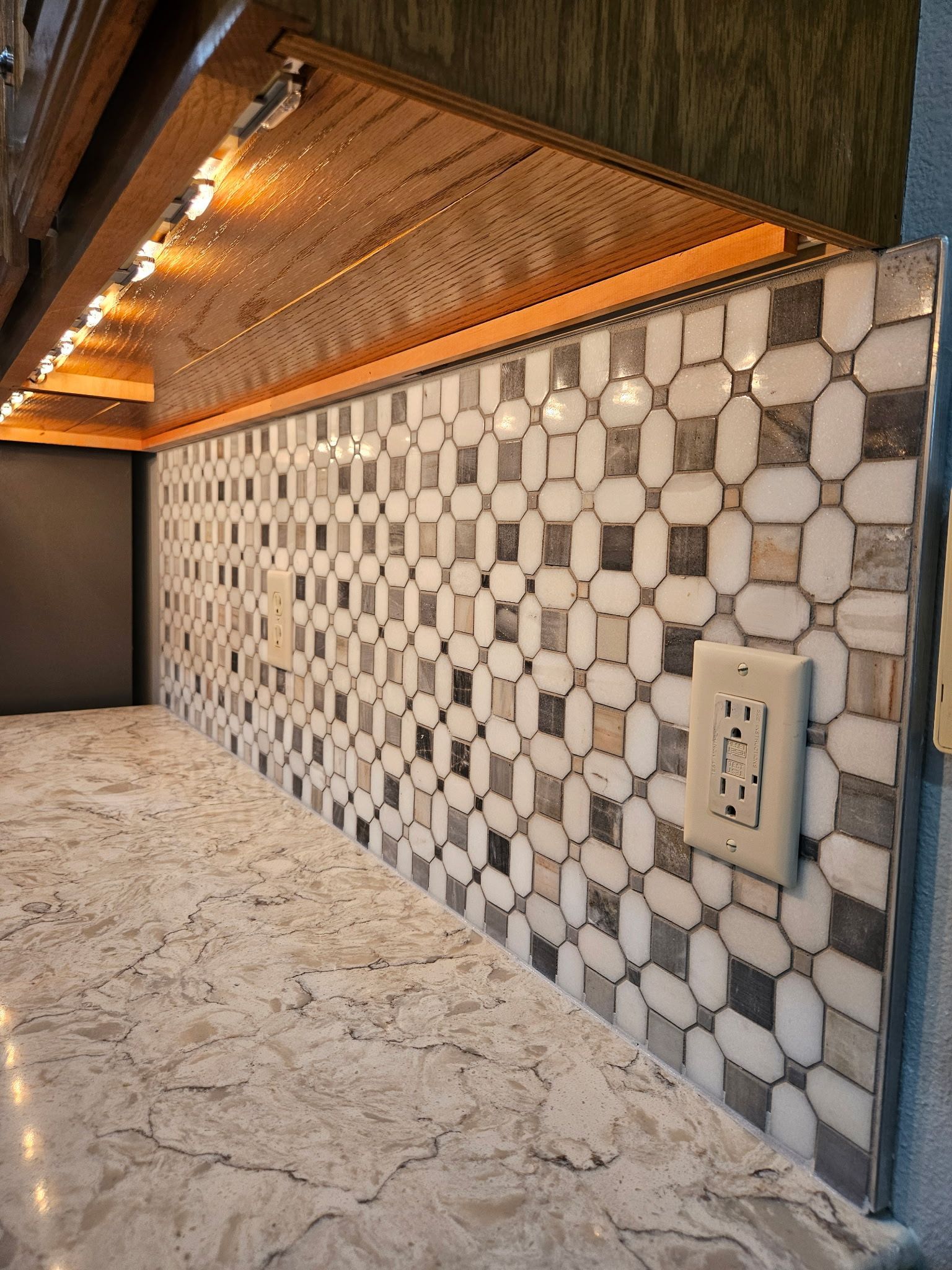 Kitchen backsplash of patterned mosaic tile with countertop and under-cabinet lighting.
