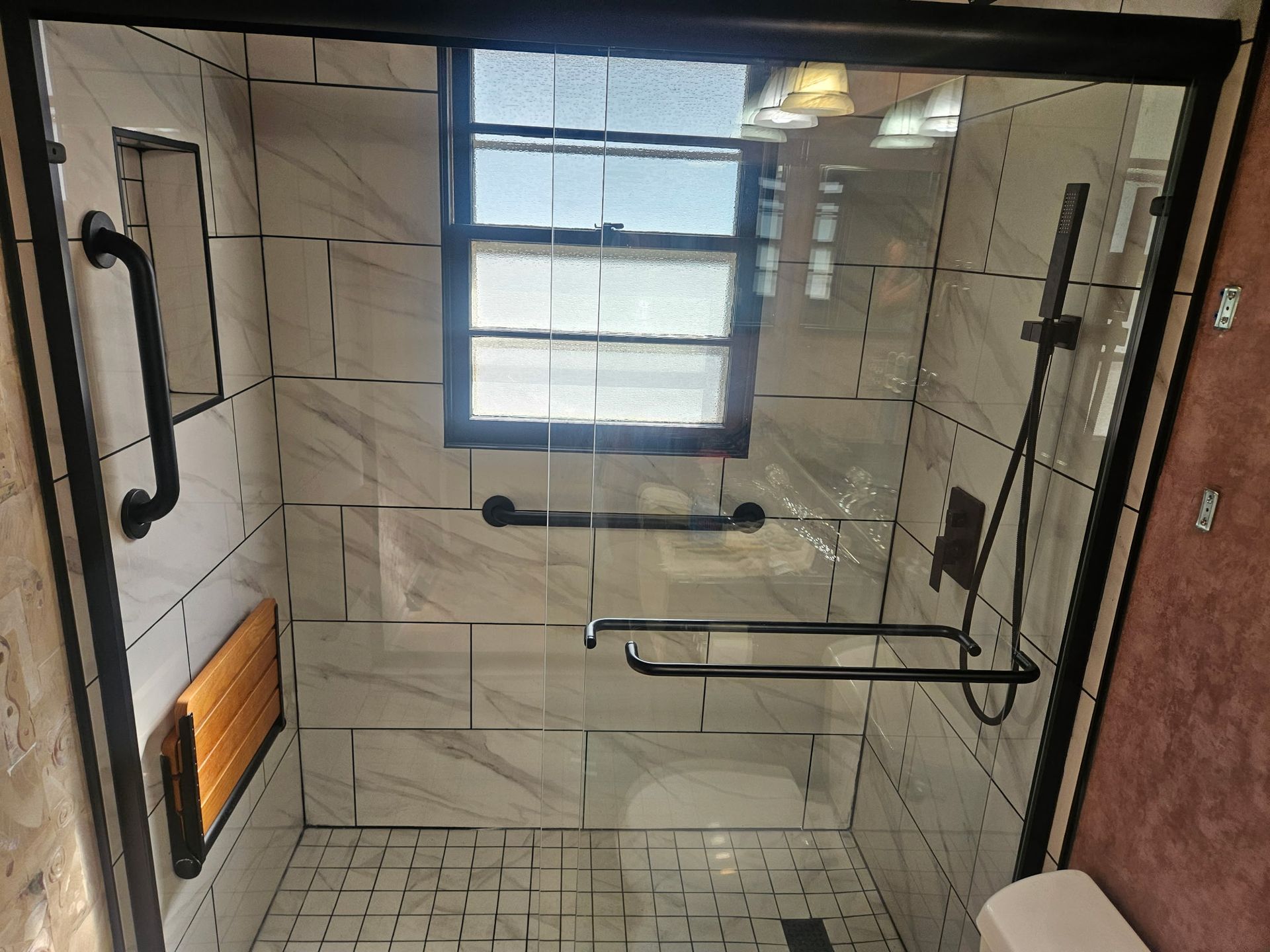 Shower stall with black fixtures and grab bars, marble-like tiles, a window, and glass doors.