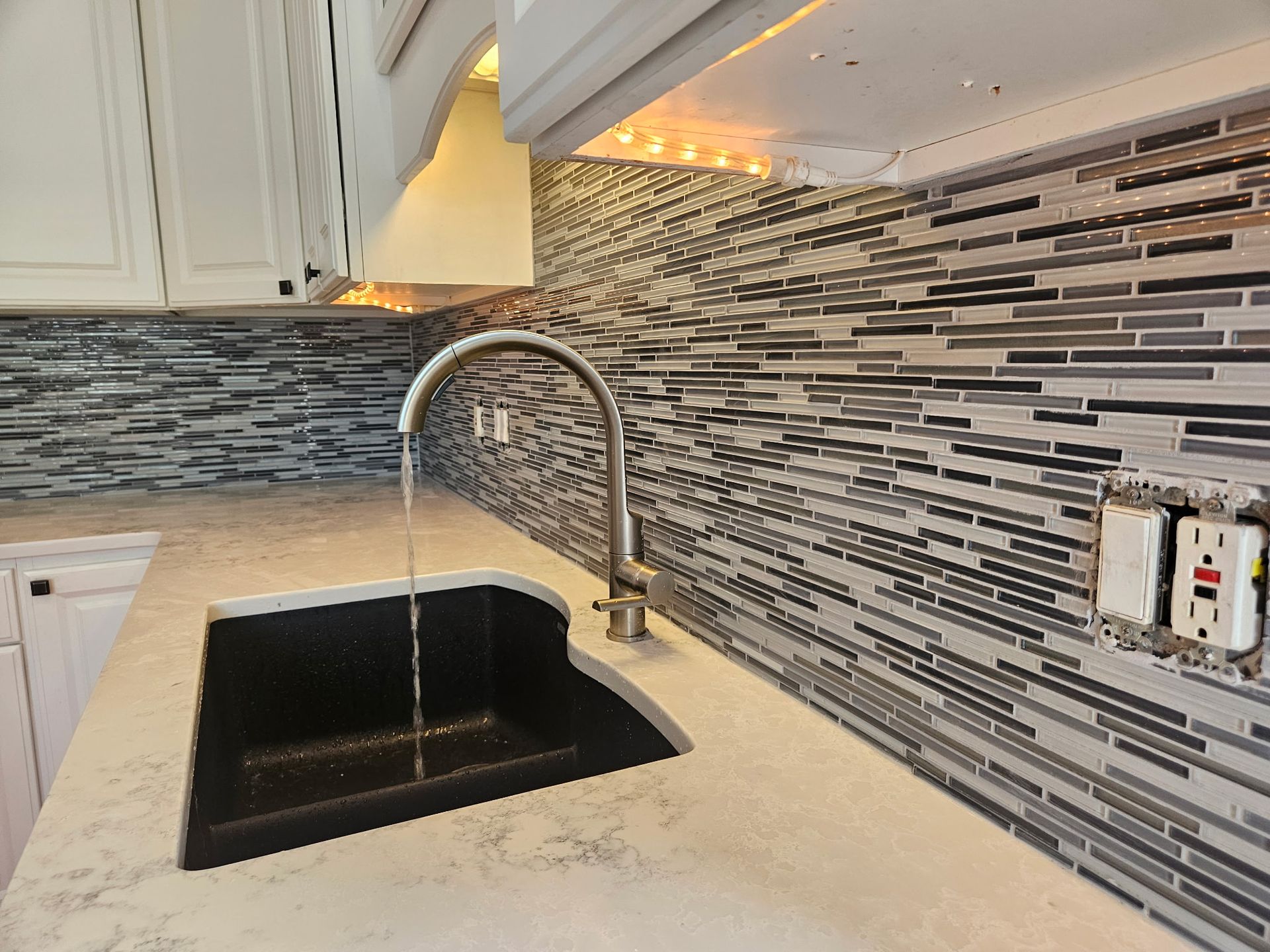 Kitchen with black sink, faucet with running water, and gray and white mosaic backsplash.