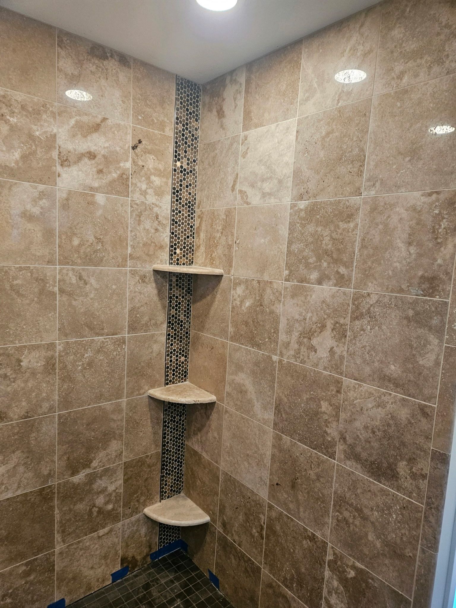 Tiled shower corner with decorative mosaic accent and built-in shelves. Earth-toned tiles and recessed lighting.